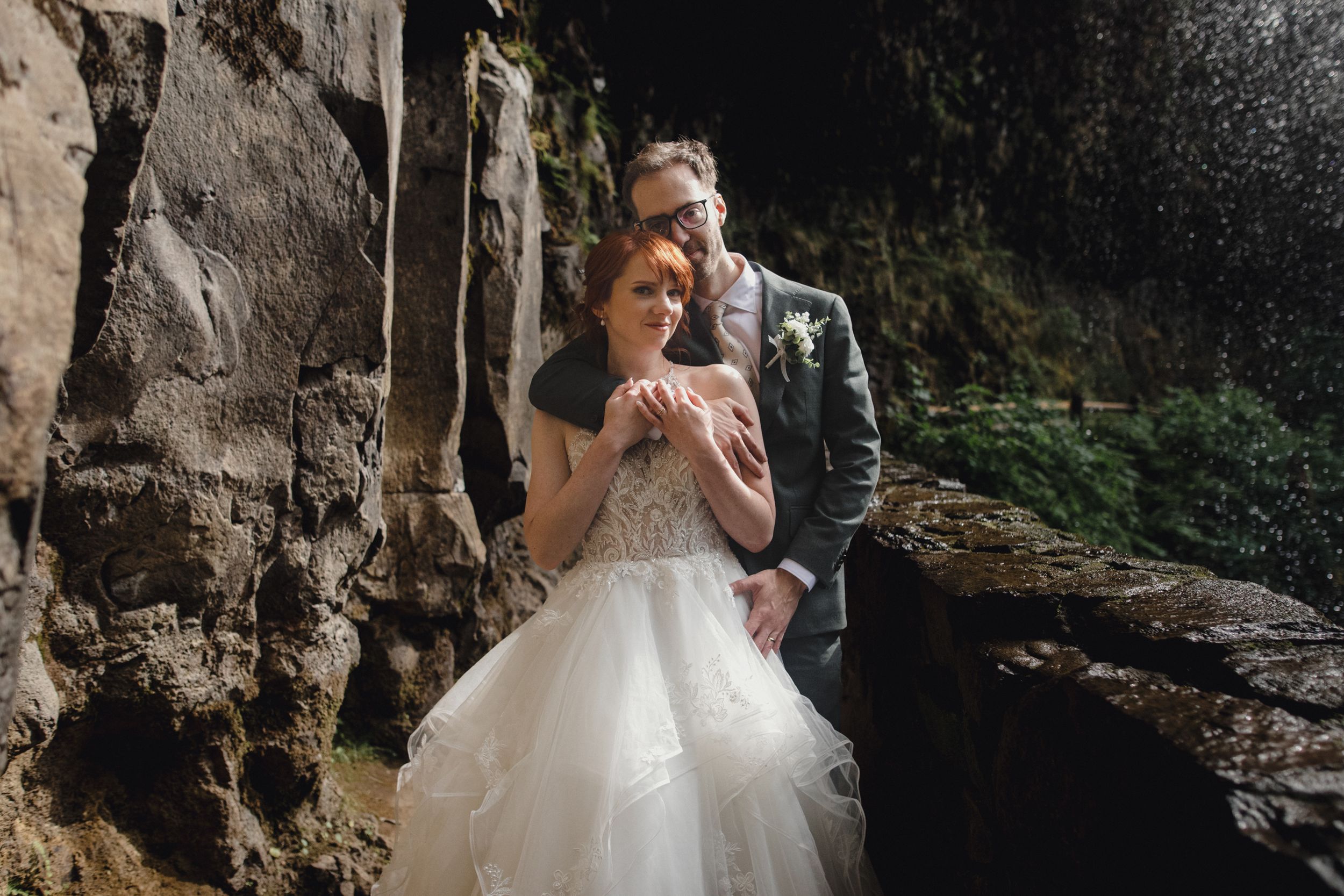 A couple in wedding attire share an intimate moment near a rocky cliff wall in a natural outdoor setting.