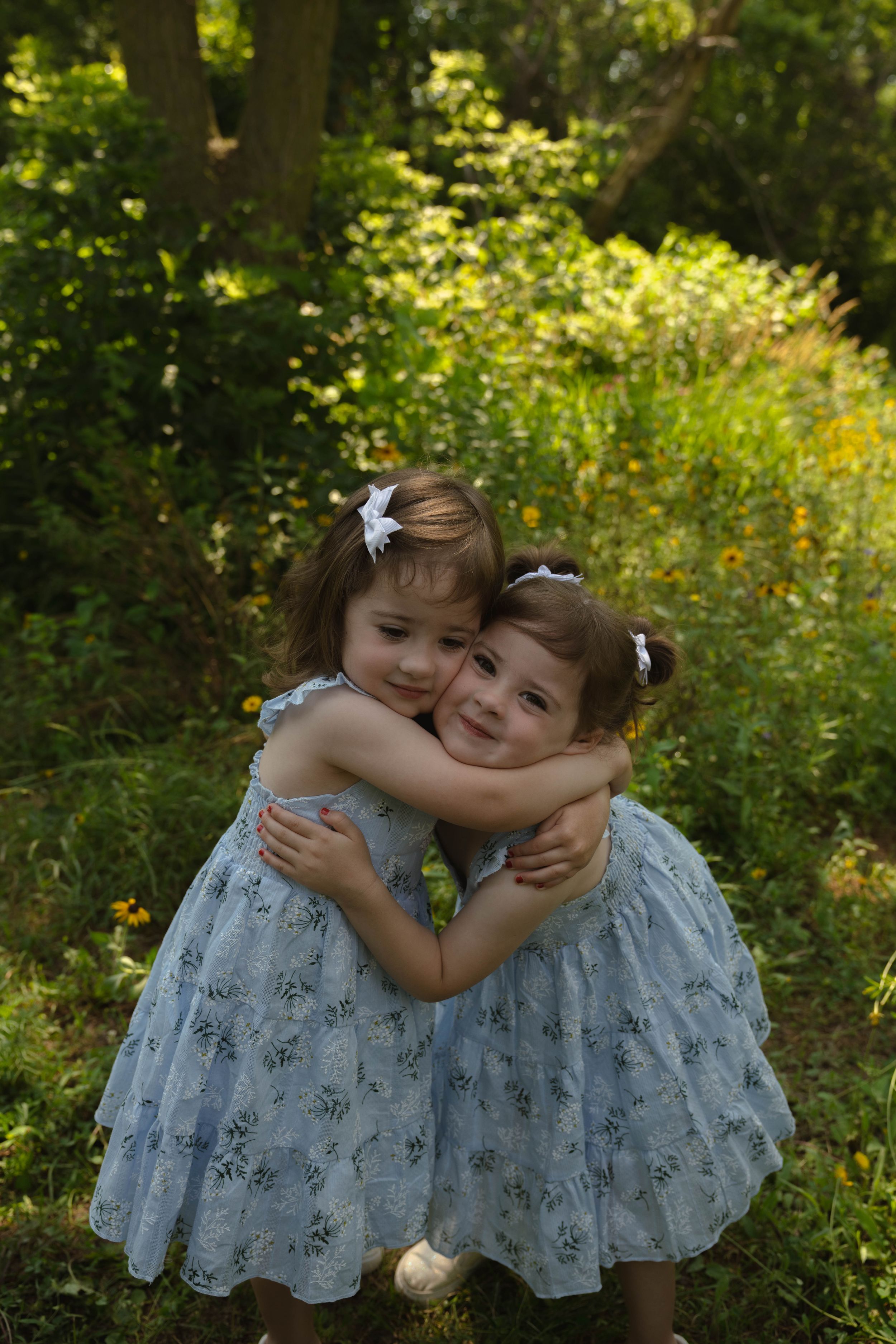 Two children in matching blue dresses hugging in a sunny garden.