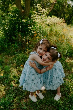 Two children in matching floral dresses share a sweet hug in a sunny garden.