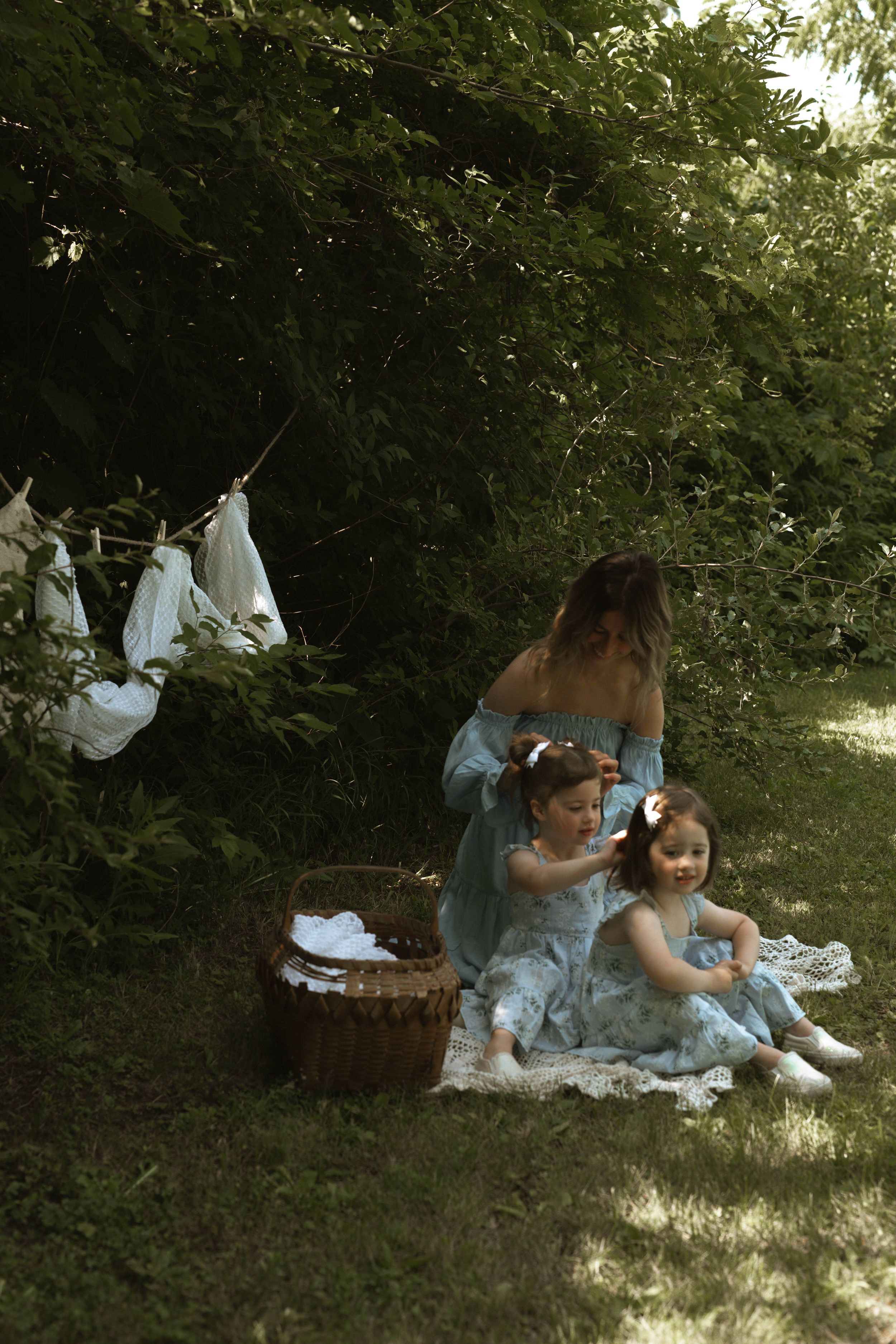 Outdoor scene with laundry line and family sitting on blanket in dappled sunlight.