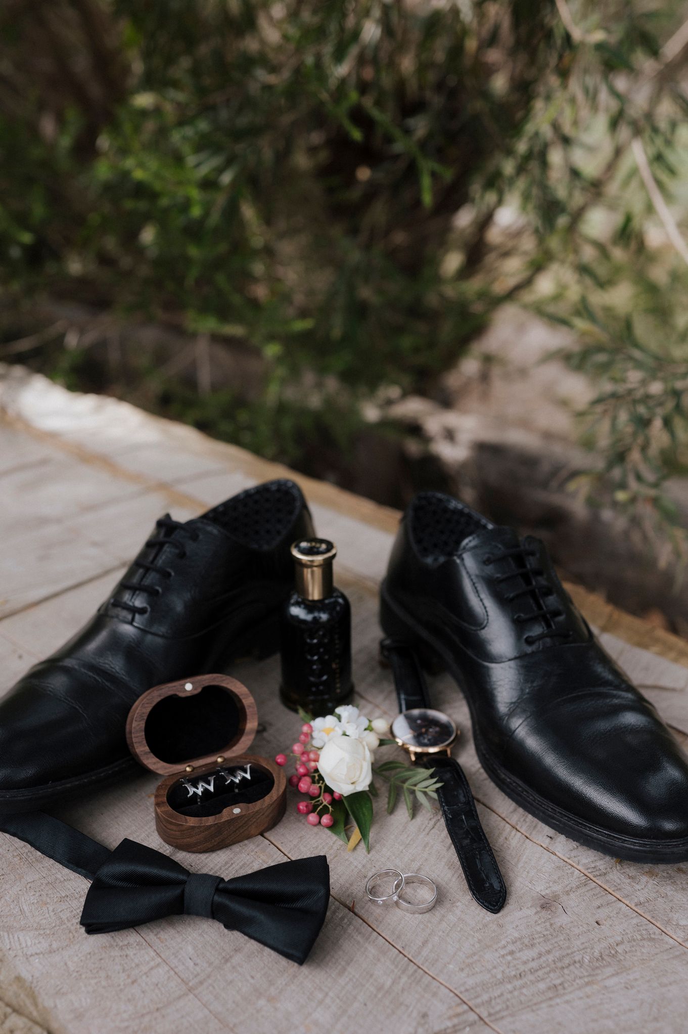 Wedding accessories including black dress shoes, bow tie, rings and boutonniere on wooden surface.
