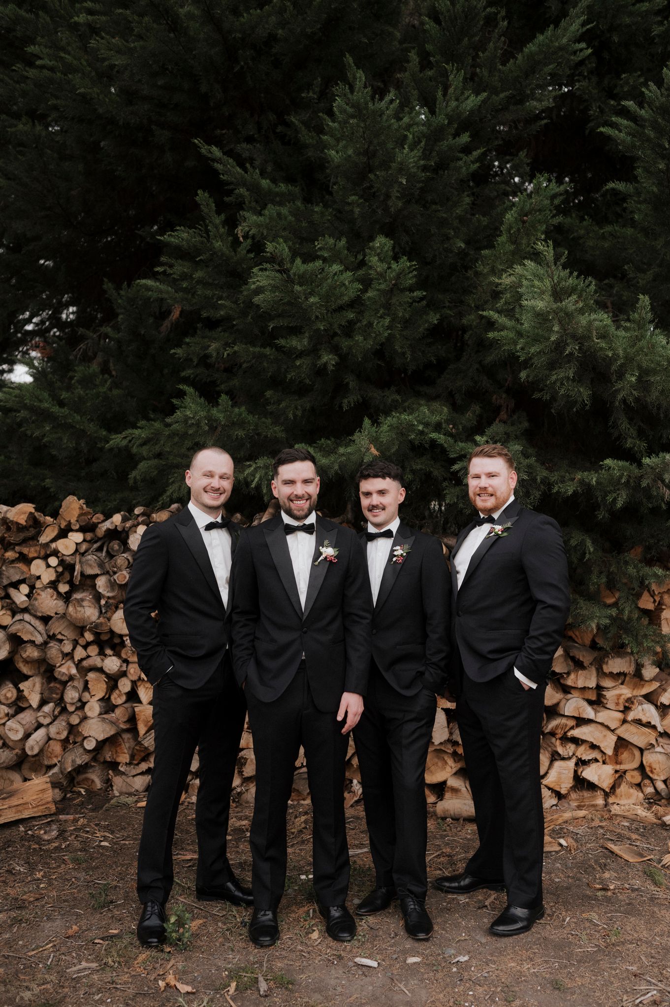 Groomsmen in black tuxedos standing together against woodpile and evergreen trees.