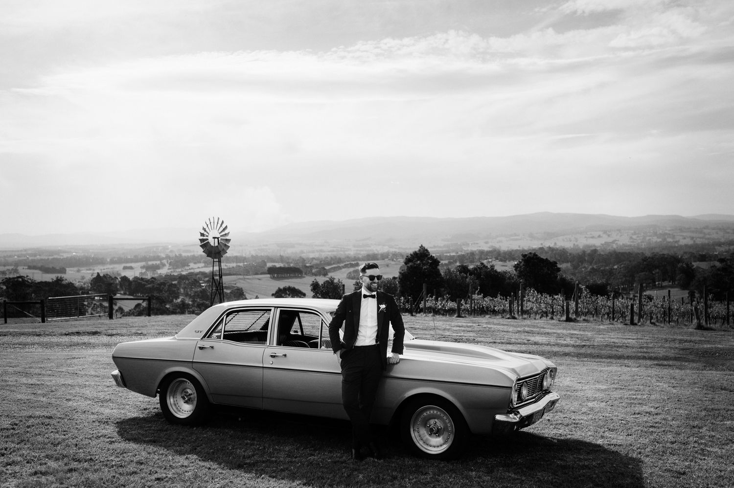 Classic vintage car with formal attire against rural landscape backdrop.