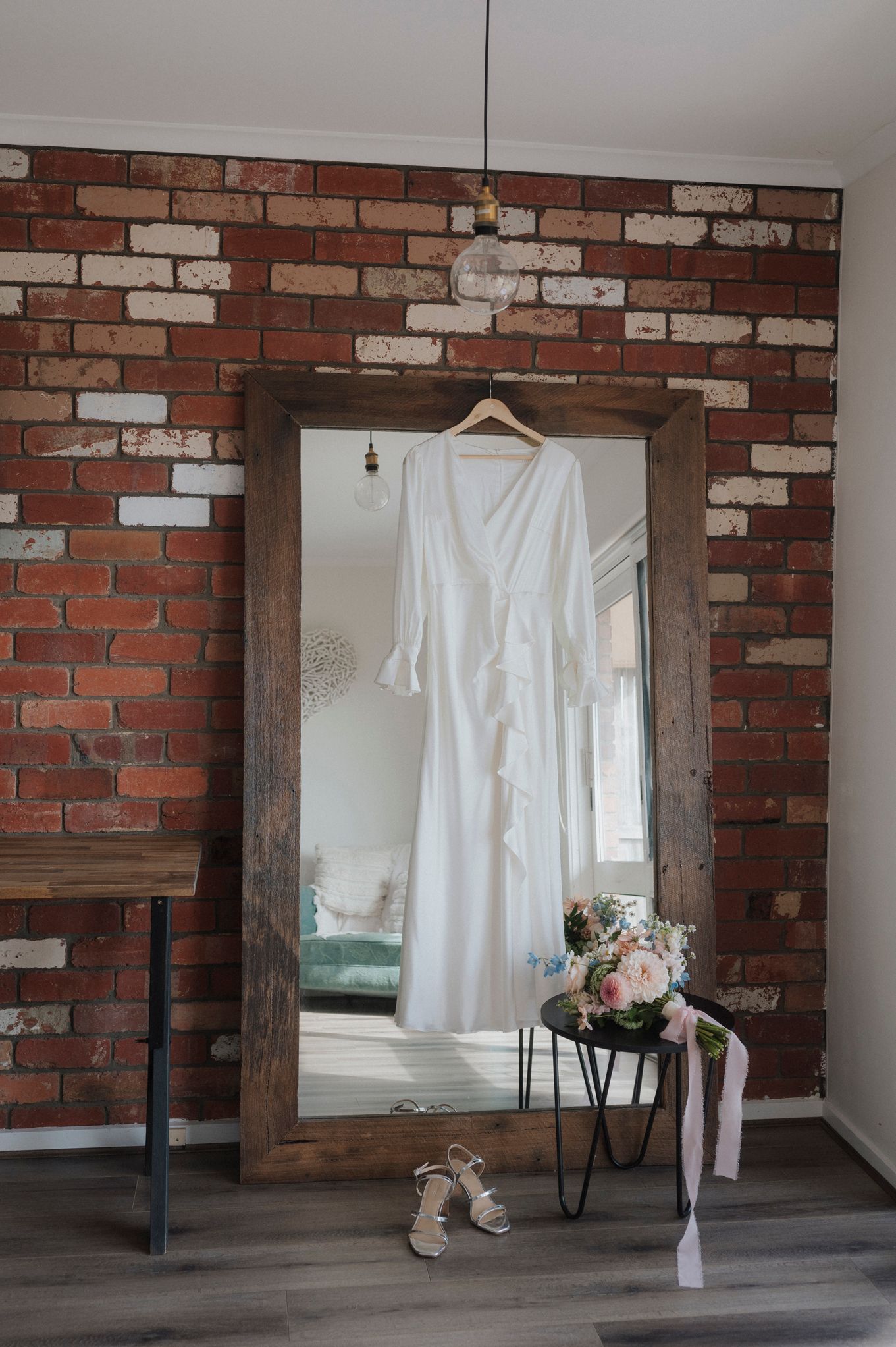 White wedding dress hanging on rustic mirror frame against exposed brick wall with floral decor.