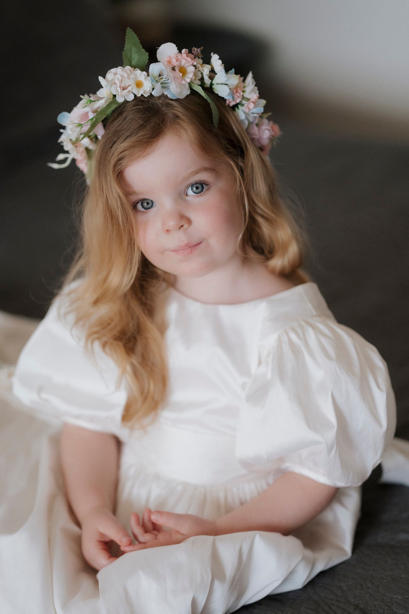 A young person in a white dress wears a floral crown with soft pink and blue flowers.