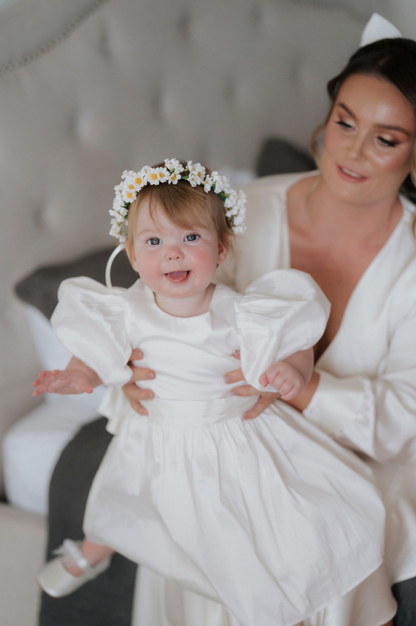 A small child in a white dress smiles while wearing a floral crown during a special occasion.