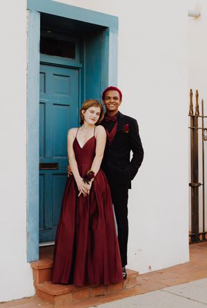 Couple in formal attire poses in front of turquoise door with burgundy gown and black suit.