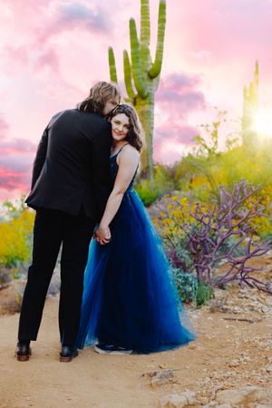 Couple in formal attire embraces on desert path with saguaro cactus at sunset.