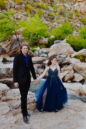 Couple in formal attire walks hand in hand through rocky desert landscape.