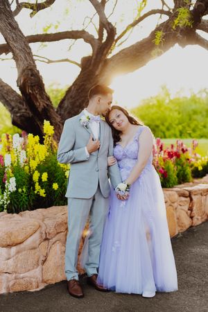 Prom couple in formal attire pose together in front of colorful spring flowers and a tree.