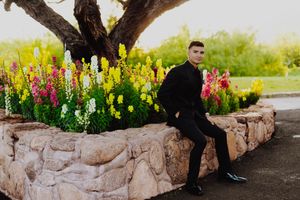 Person in black formal wear sits on stone wall with colorful snapdragons garden behind.
