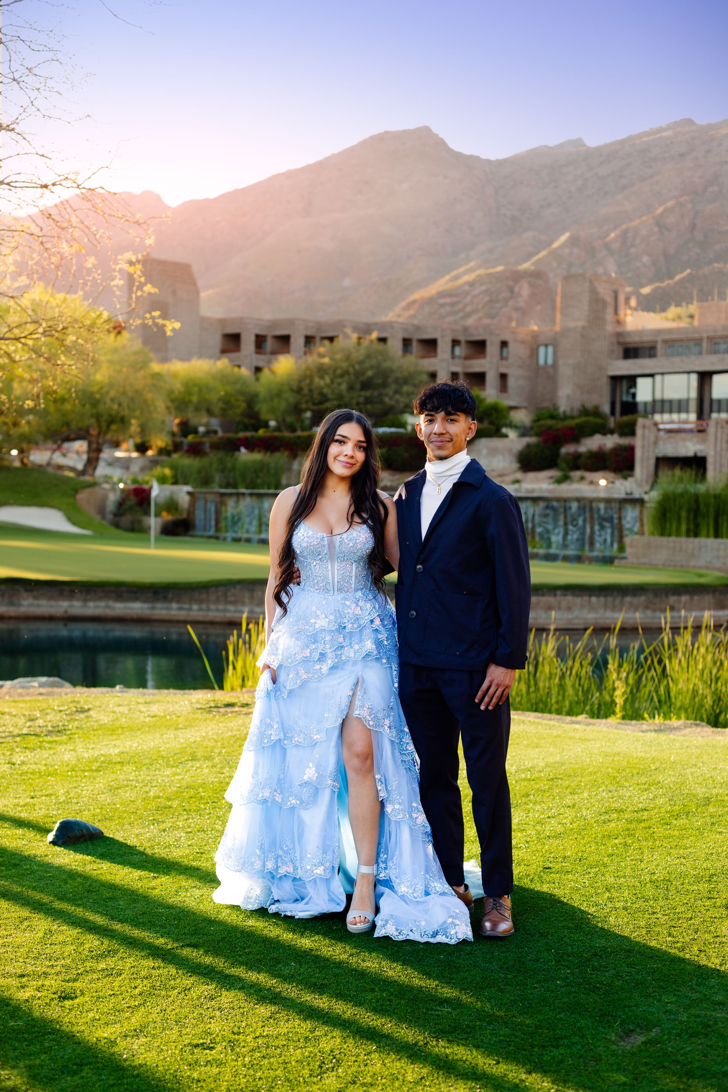 Couple in formal attire posing on golf course with mountain backdrop at sunset.