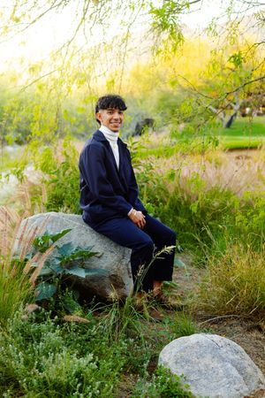 A person in a navy suit sits on a rock surrounded by golden natural foliage at sunset.