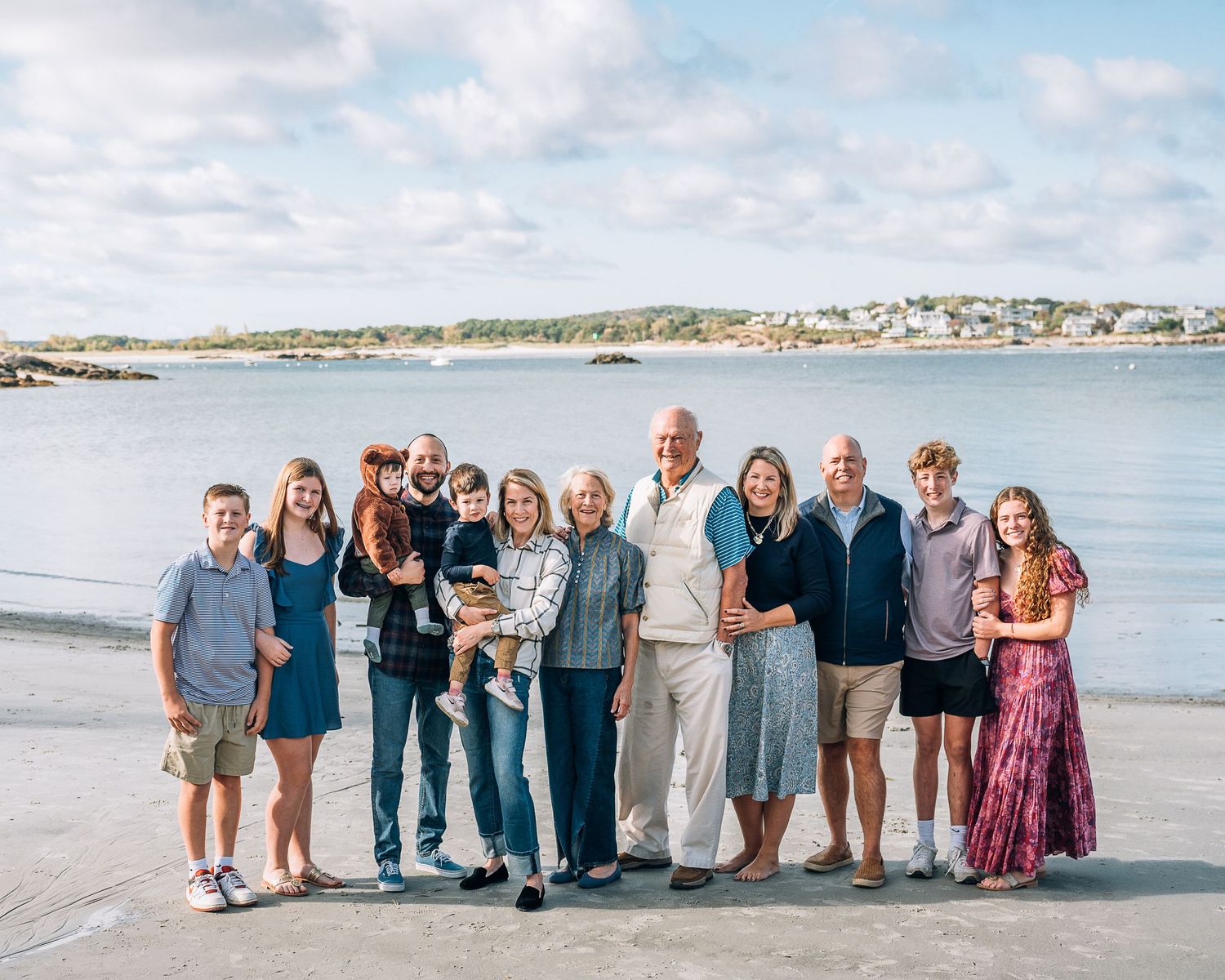 A large family group poses together on a beach with coastal homes visible in the background.
