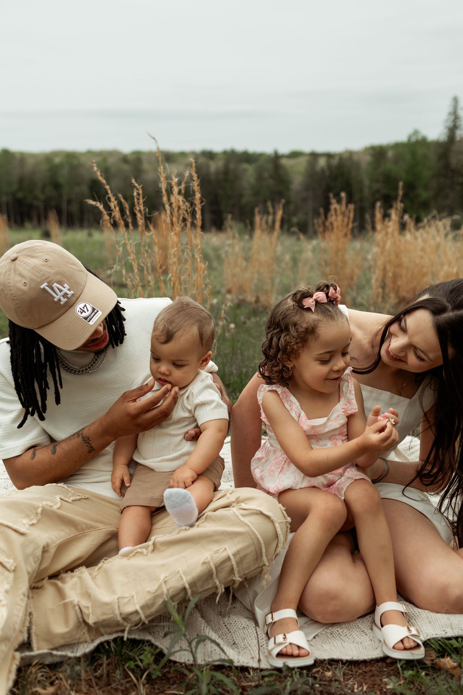 A couple in white outfits share a joyful moment with their twin babies outdoors in a wooded setting.