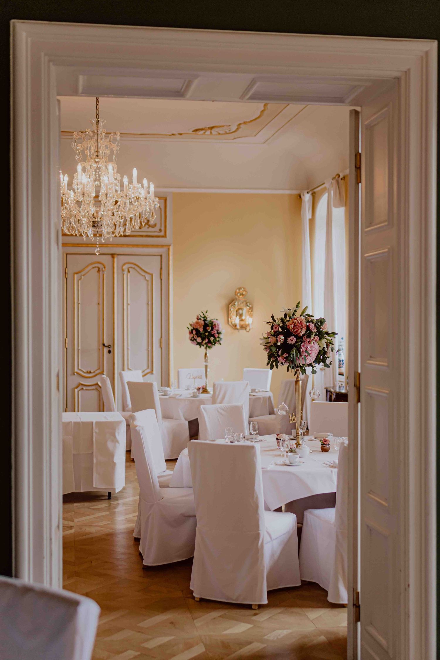 Elegant white ballroom with crystal chandeliers, ornate wall moldings and white-slipcovered chairs arranged for a formal event.