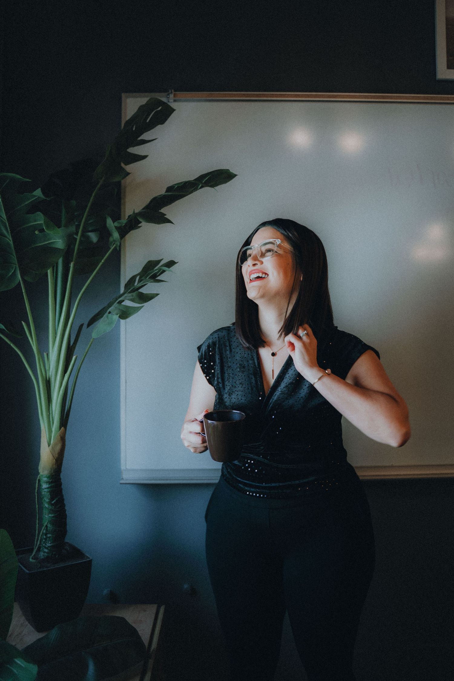 A person in a black dress poses dramatically against a moody dark wall with a large indoor plant.