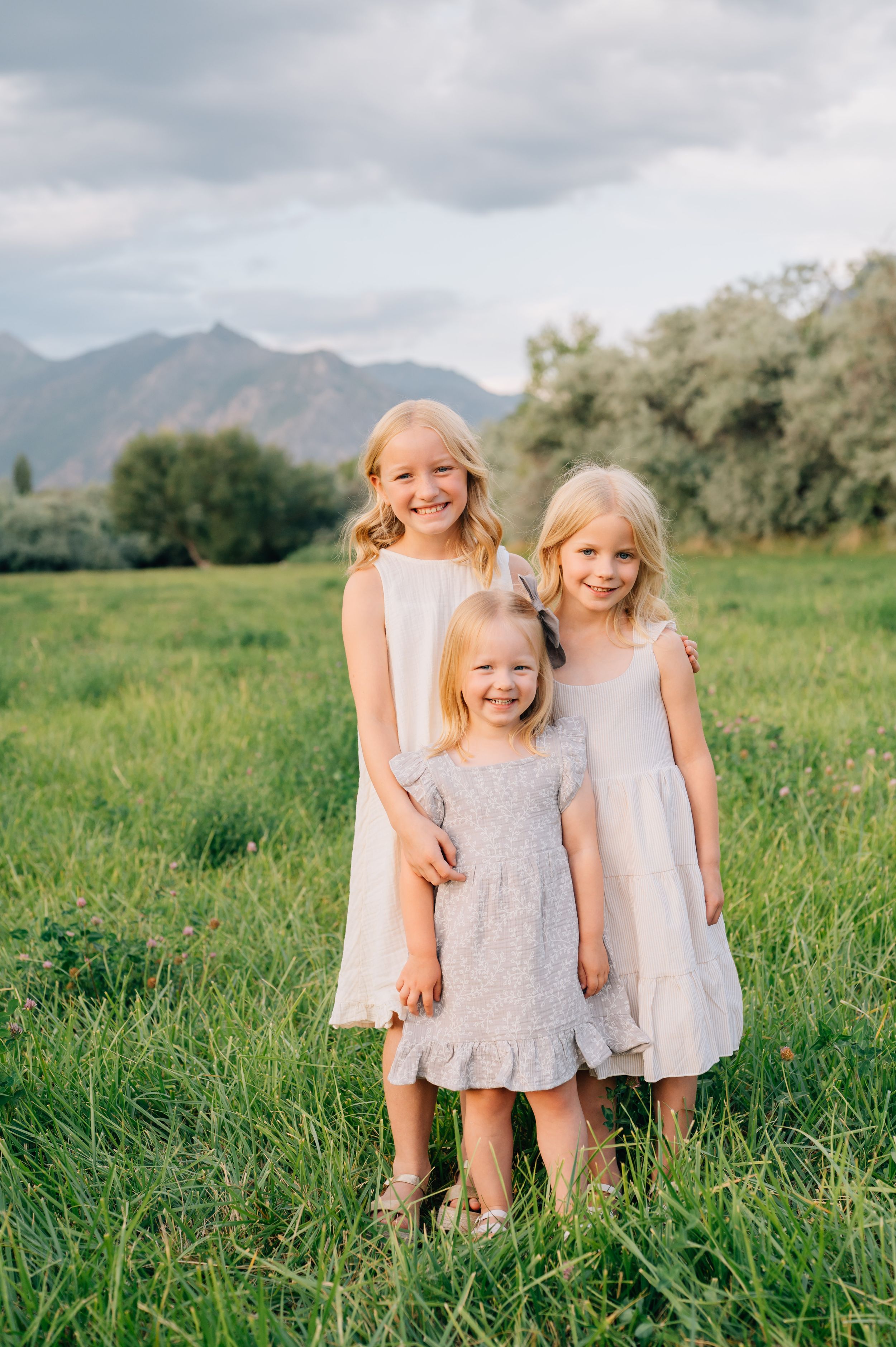 Three young children wearing light colored dresses stand together in a grassy meadow with mountains in the background.