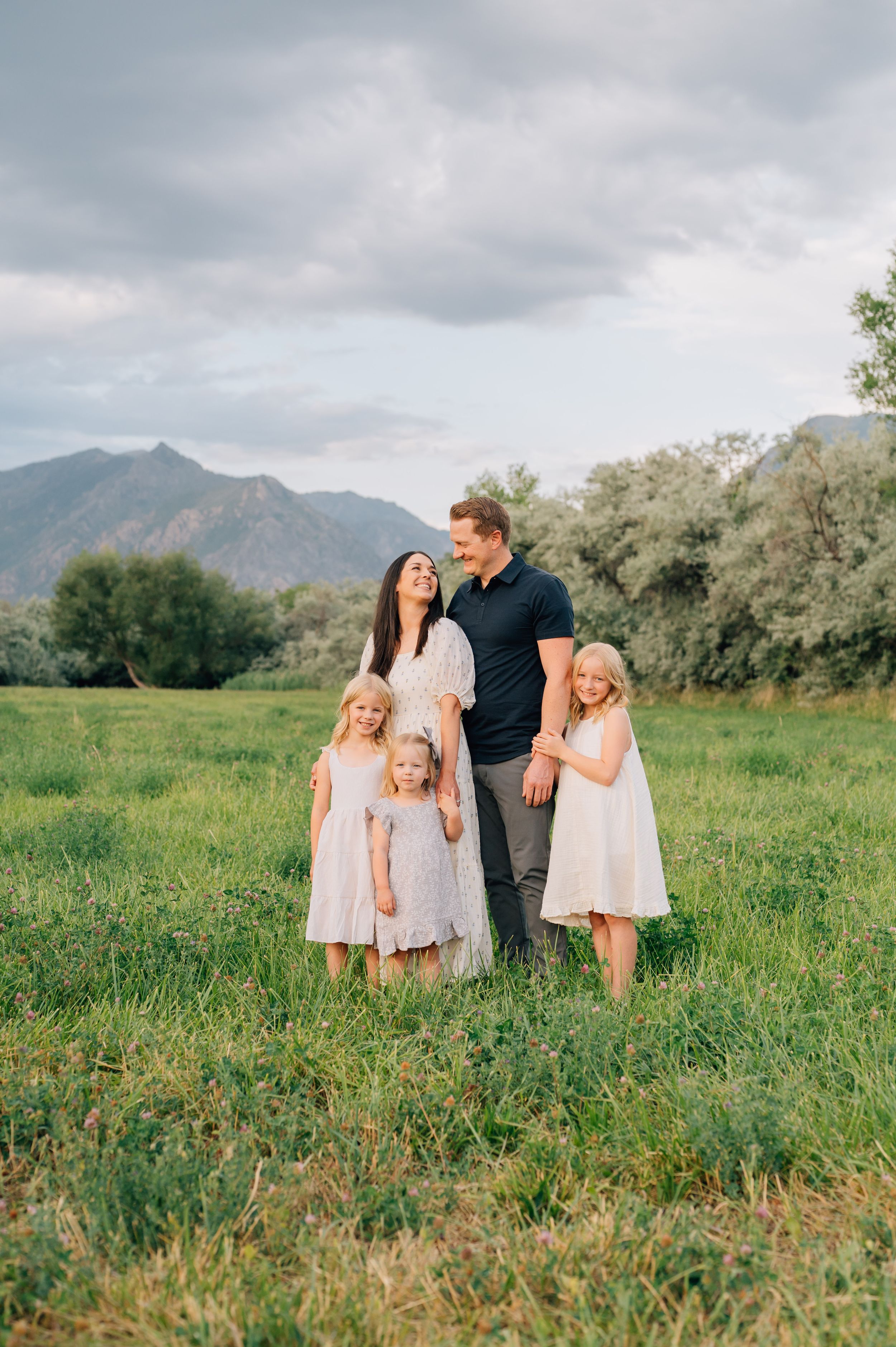 Casual family portrait in a green meadow with scenic mountain backdrop on an overcast day.