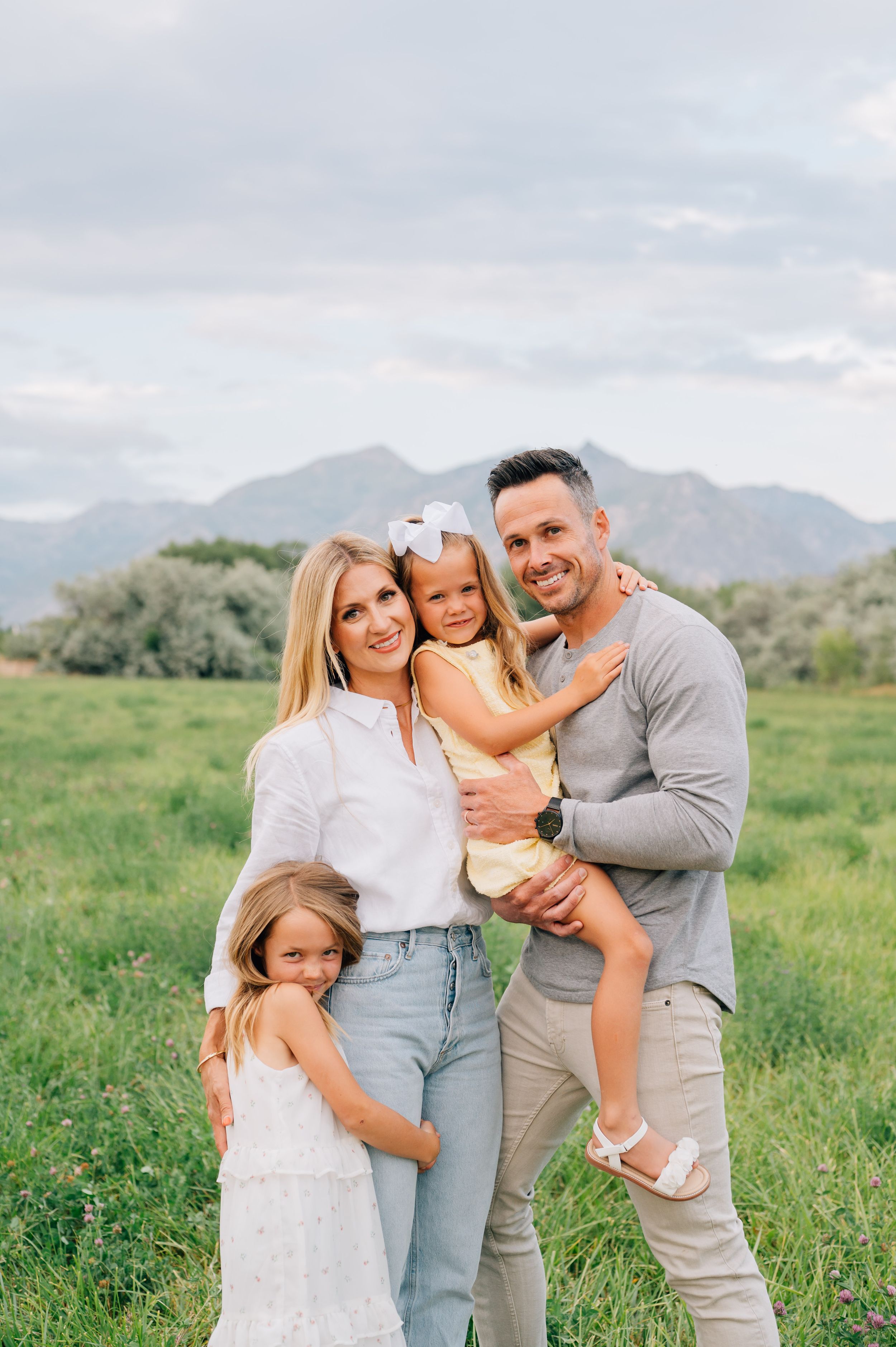 A family wearing light-colored clothing poses together in a sunny green meadow with mountains in the background.