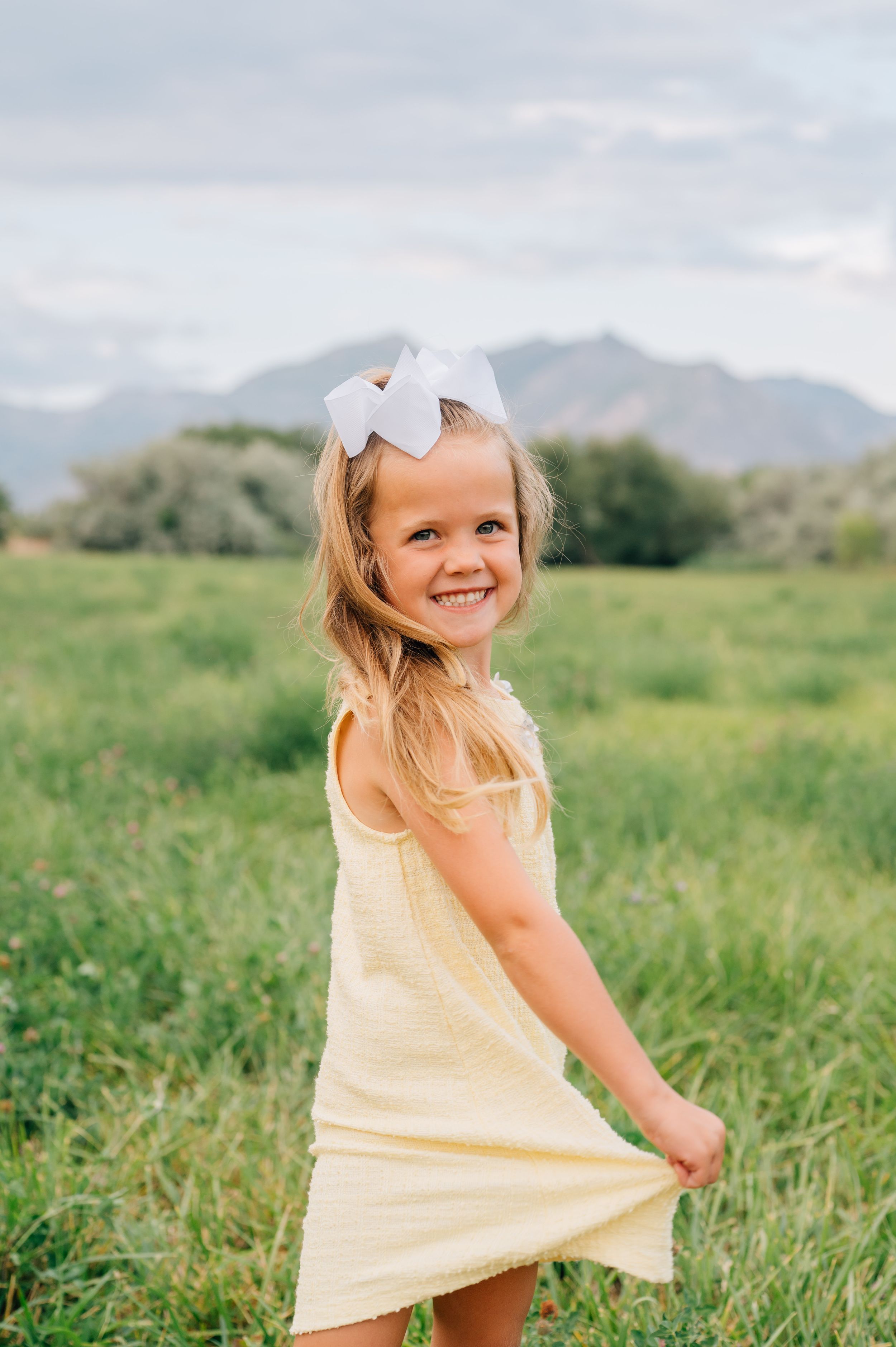 A young child in a yellow dress twirls playfully in a green field with mountains in the background.