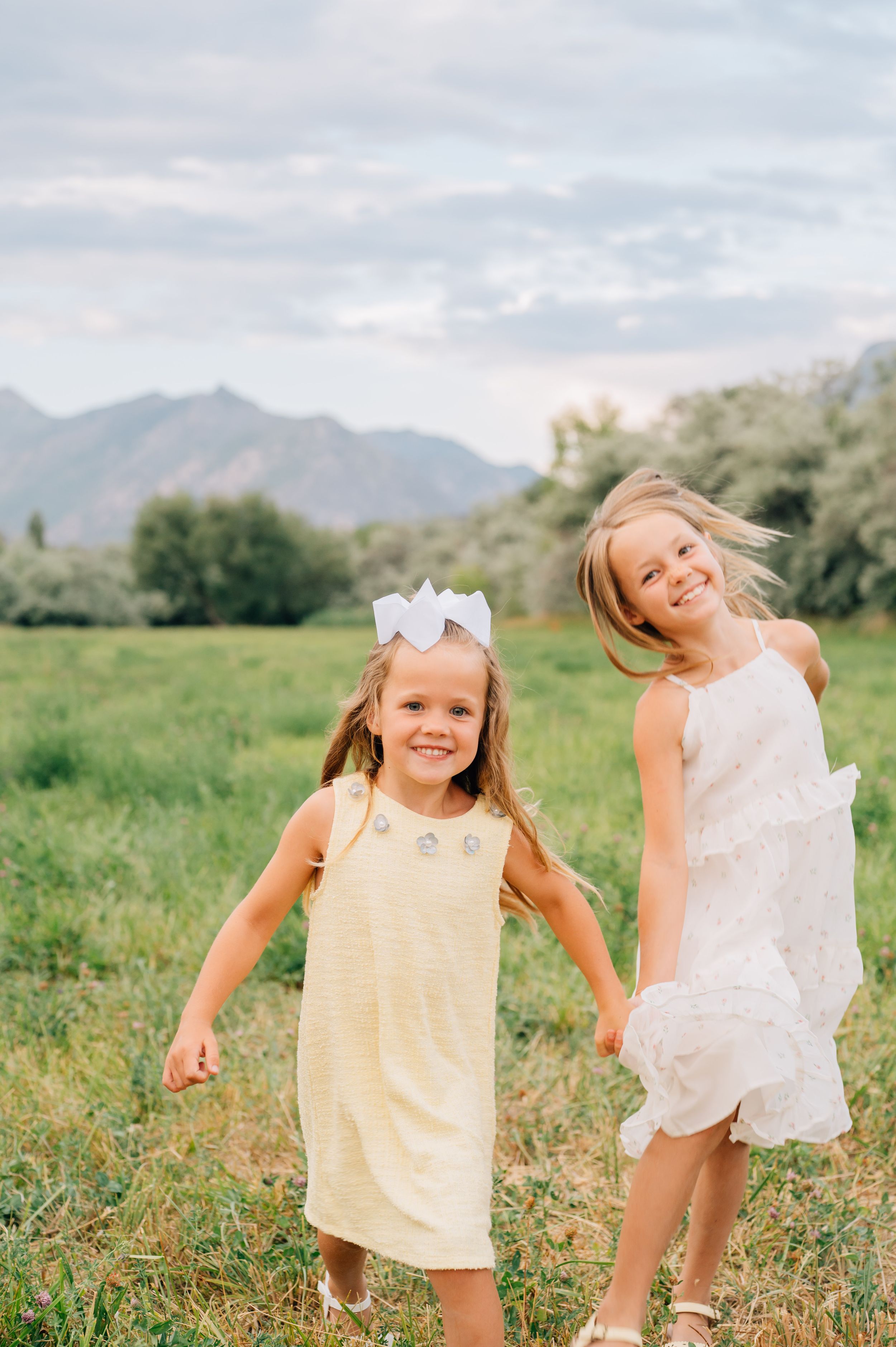 Two siblings in light summer outfits playing joyfully in a mountain meadow with green grass and clear skies.