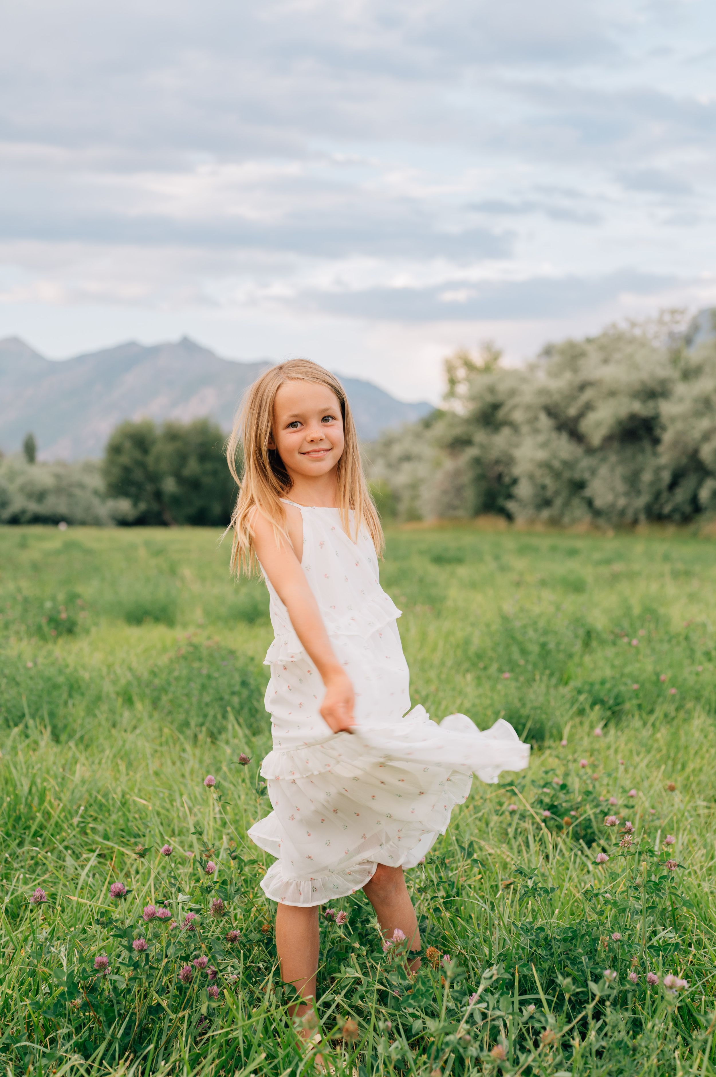 A child in a white summer dress twirls and dances joyfully in a green mountain meadow in three sequential photos.