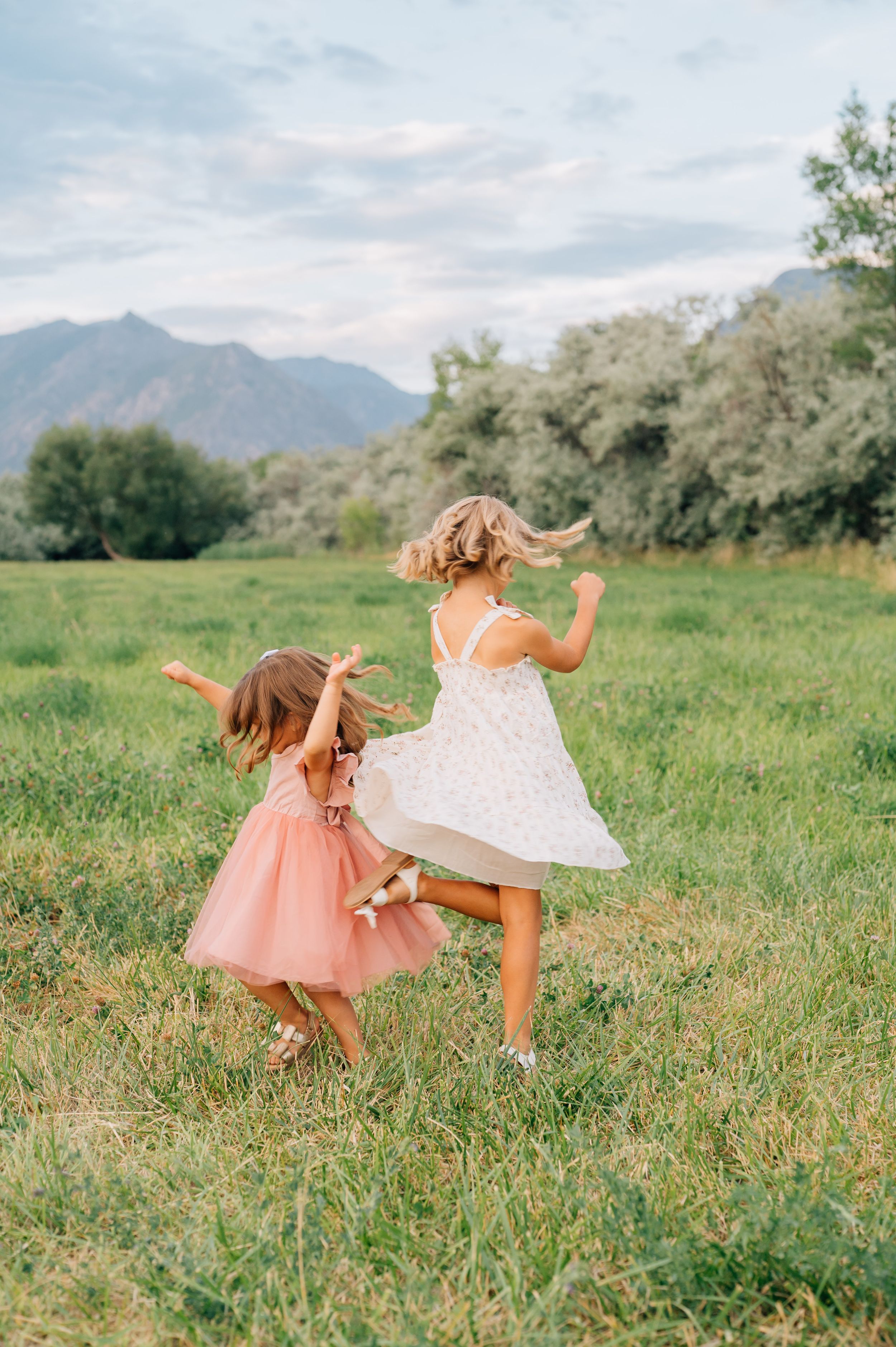 Two children in dresses dance and twirl joyfully in a green meadow with mountains in the distance.