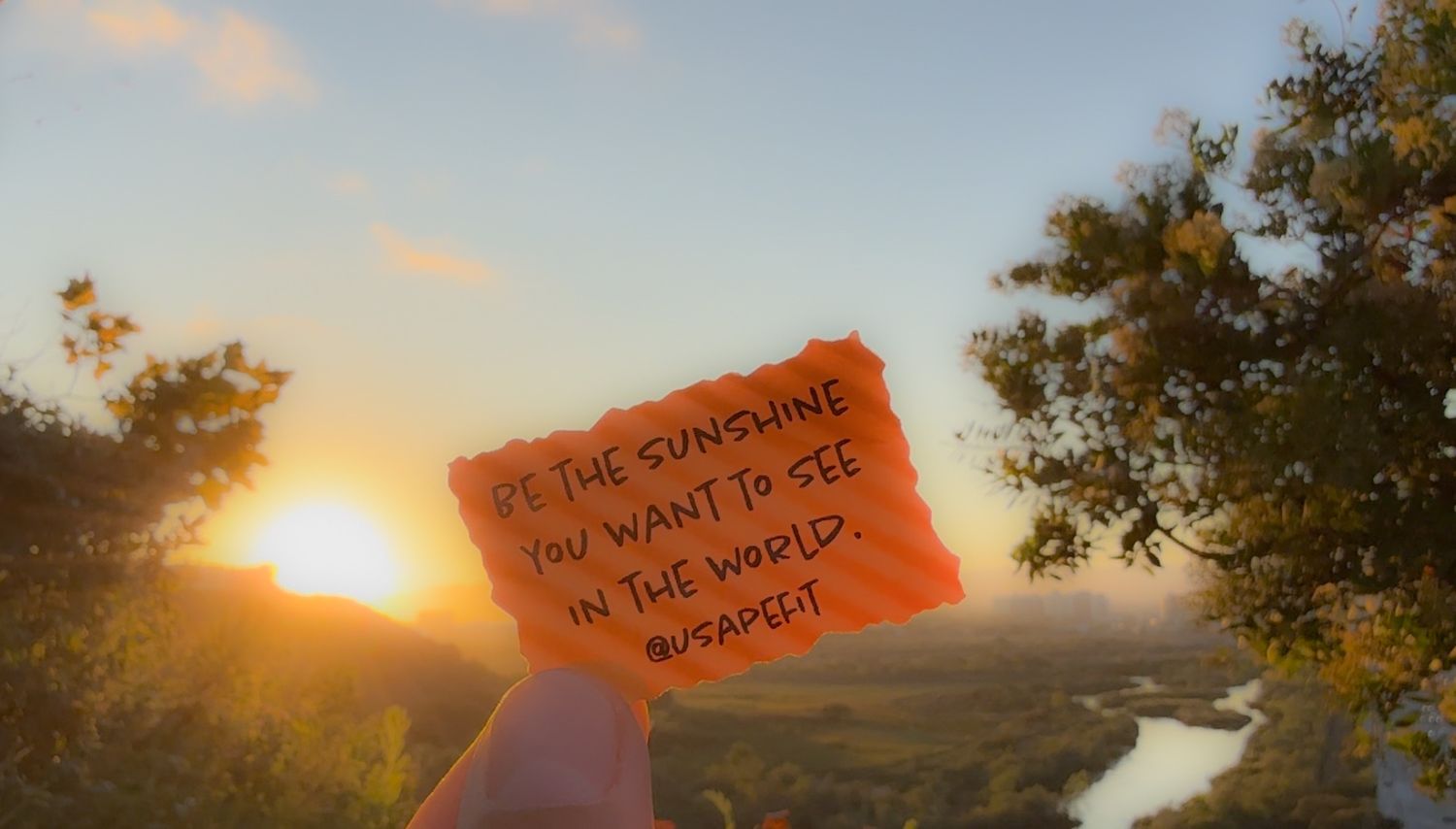 Hand holding orange note with inspirational text against sunset landscape with winding river and trees.