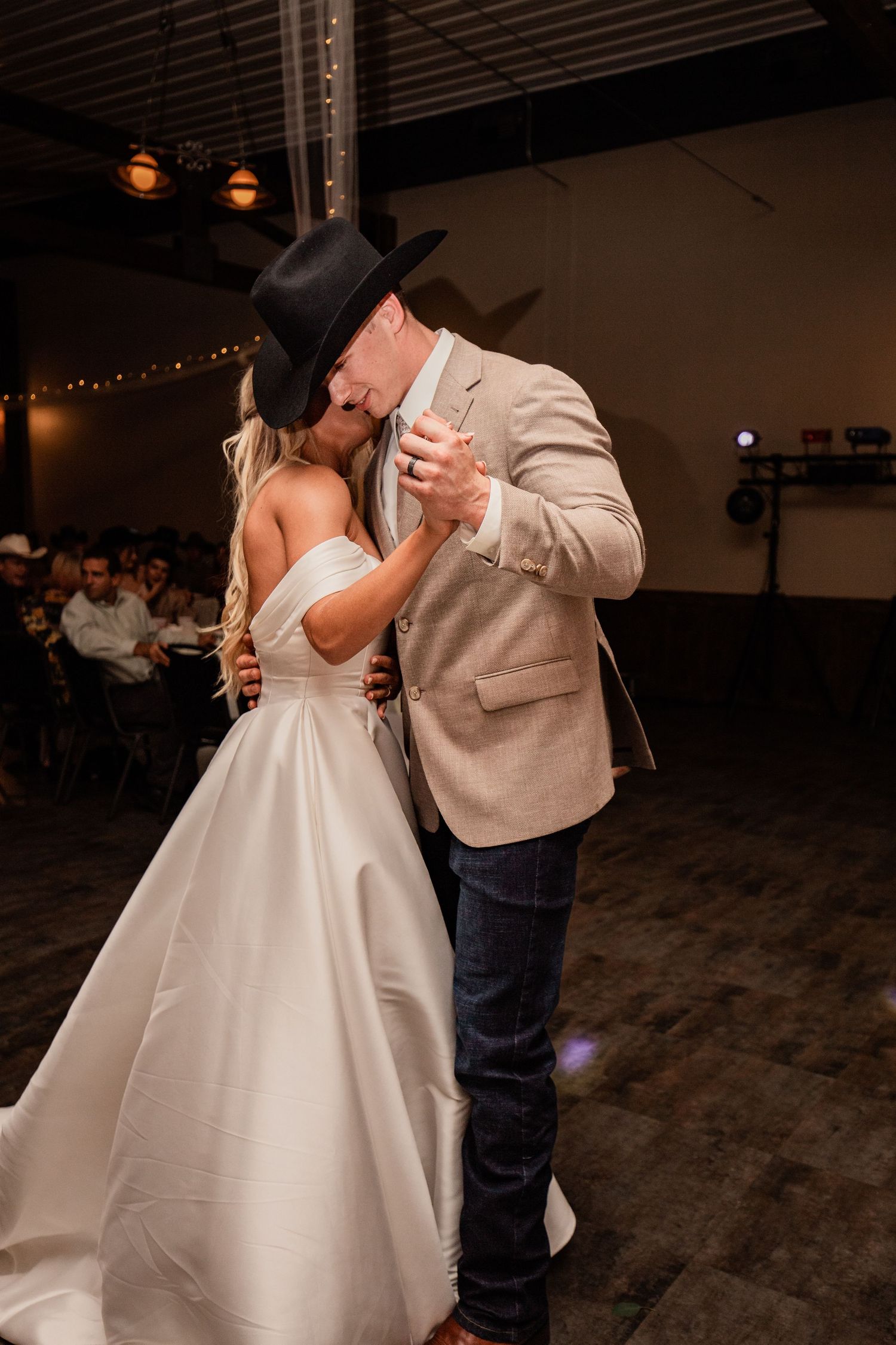 A bride in a white wedding dress dances with a partner wearing a beige blazer and black cowboy hat at a dimly lit reception.