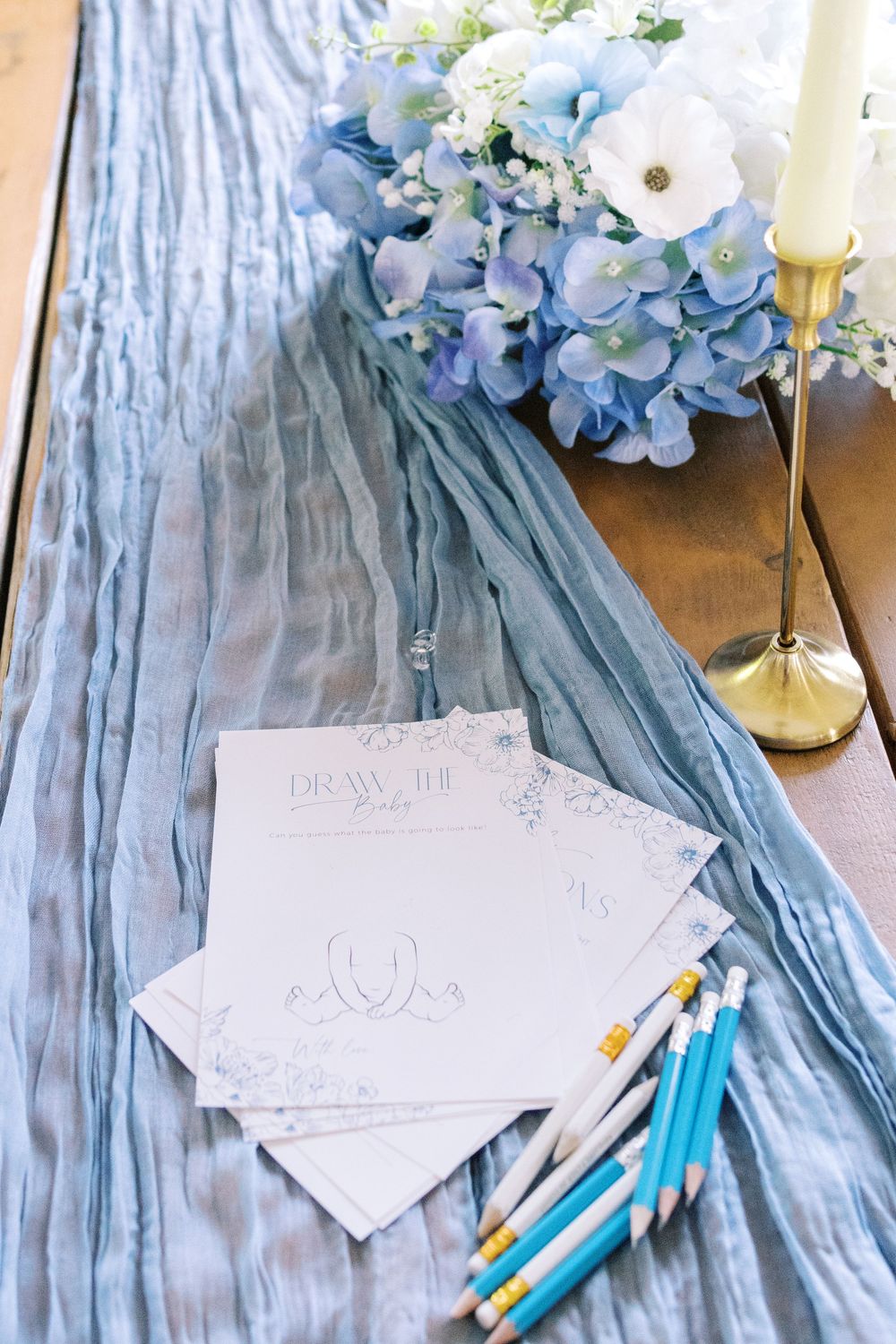 Draw the Baby game cards with blue pencils beside blue hydrangeas and candle on a blue table runner.