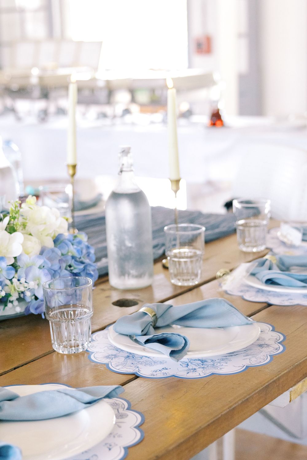 Close-up of a blue and white table setting featuring scalloped placemats, folded blue napkins, crystal glasses, and gold candlesticks.