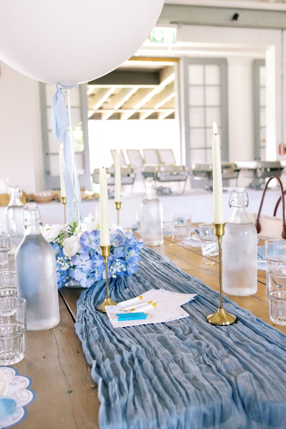 Baby shower table decor featuring a blue crinkled table runner, white balloon, gold candleholders, and blue hydrangea floral arrangement.