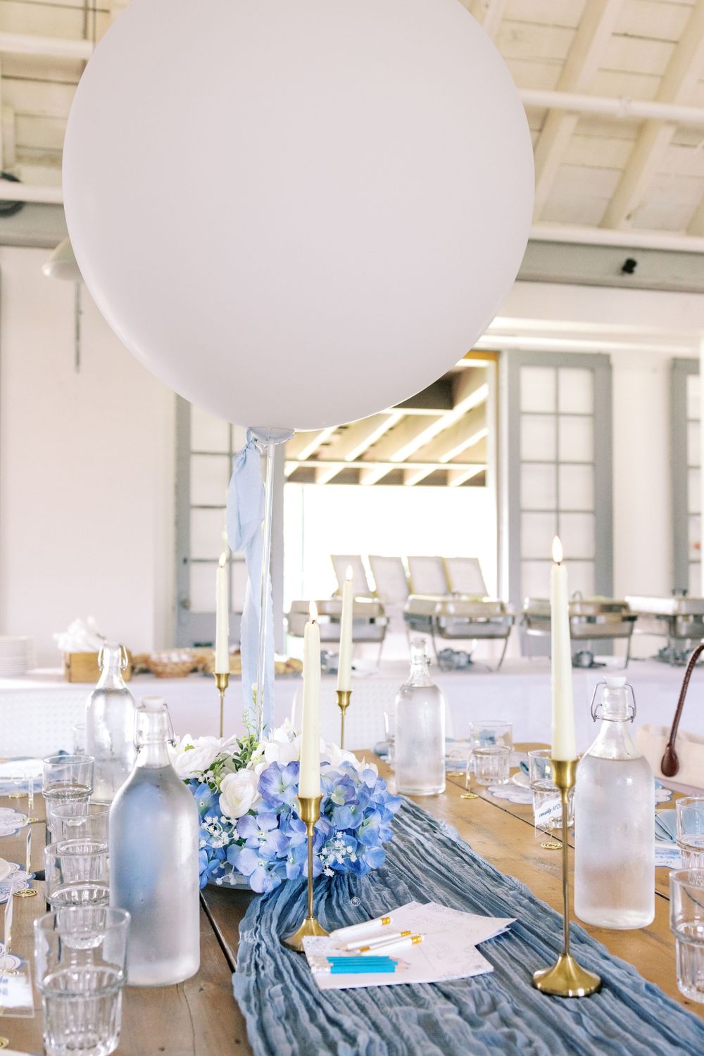 Rustic wooden table with blue hydrangea centerpiece, white balloon, gold candlesticks, and a blue gauze table runner for a party.