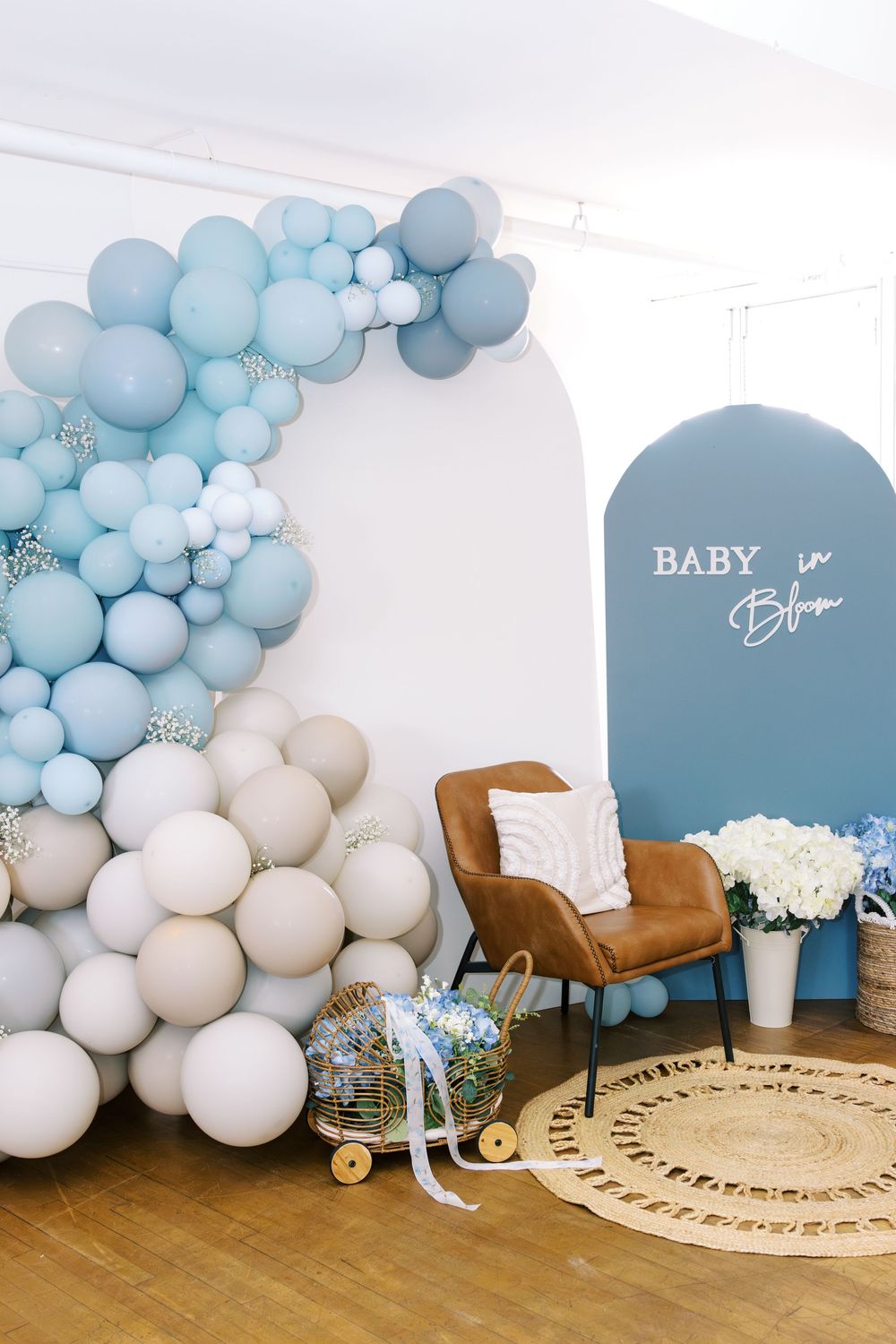 Blue and white balloon arch with beige balloons, brown leather chair, and Baby in Bloom sign at a baby shower.