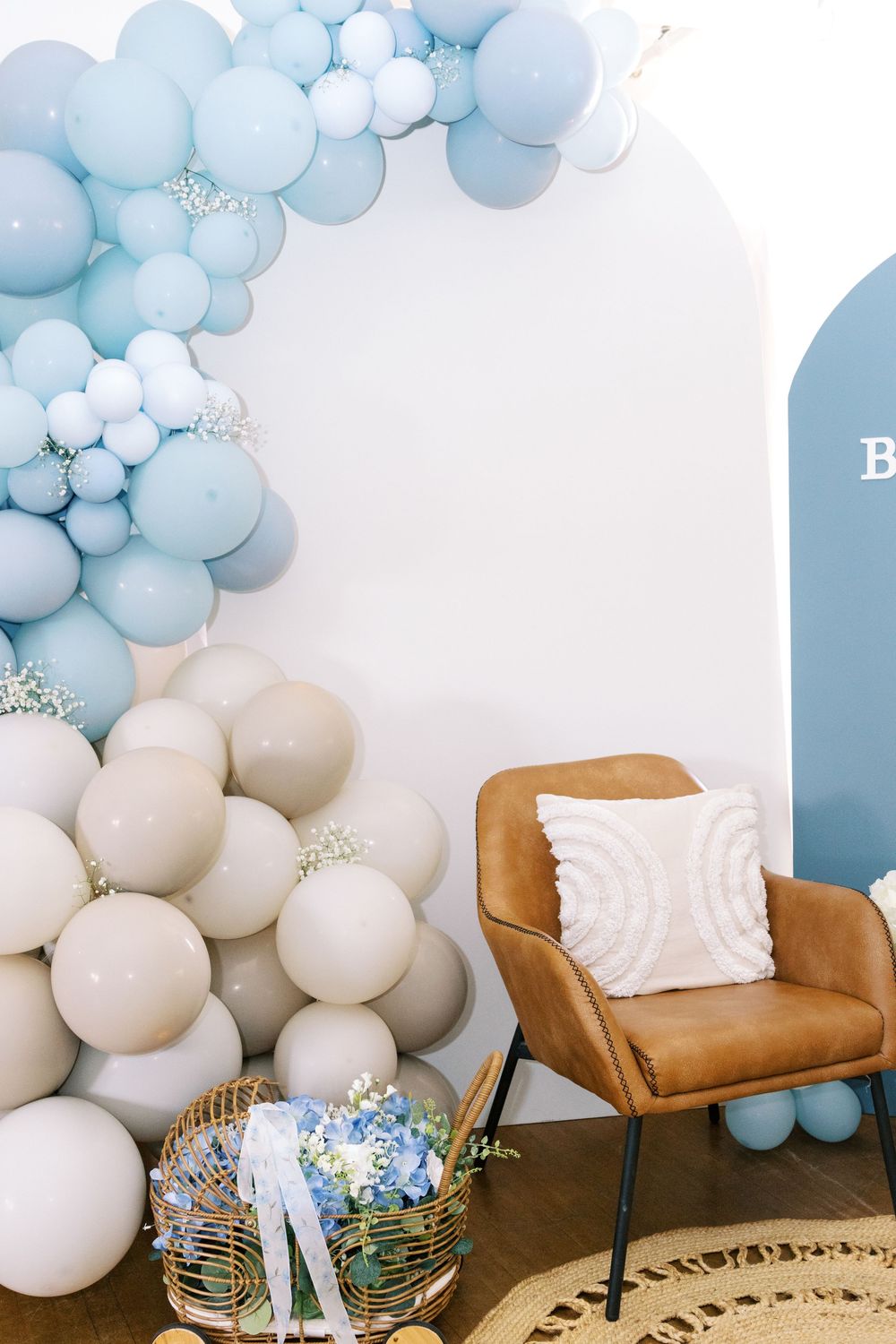Blue and beige balloon garland arch with brown leather chair and floral basket at a baby shower.