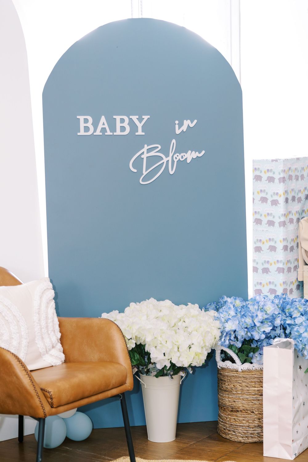 Blue arched backdrop with Baby in Bloom sign, white and blue hydrangeas, and brown leather chair.