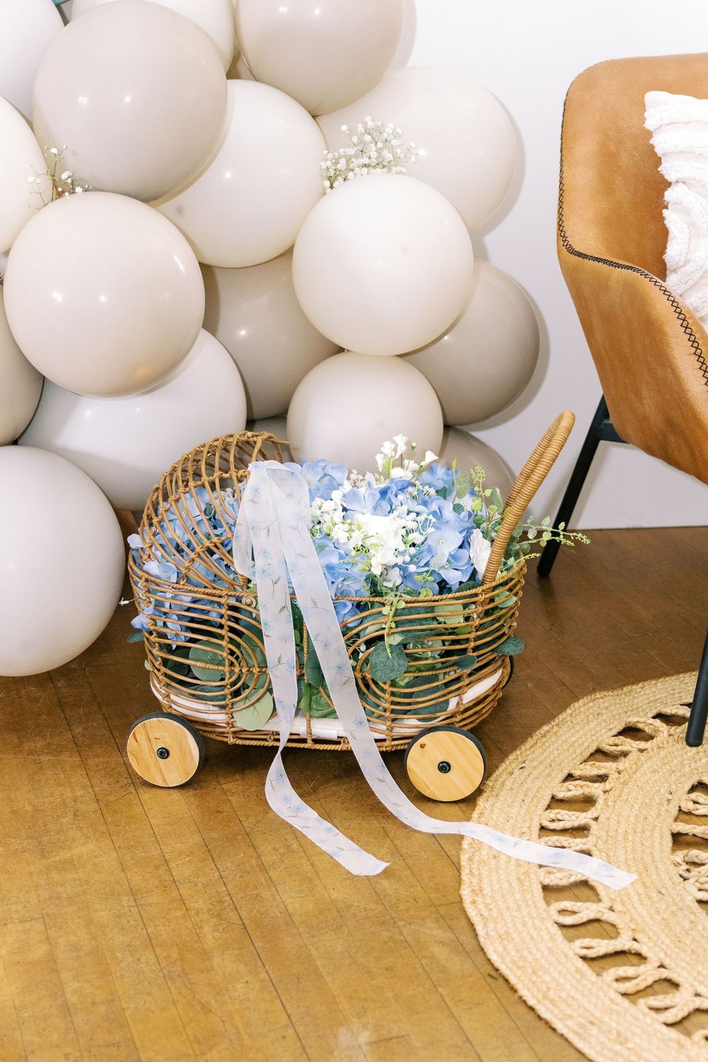 Wicker pram filled with blue hydrangeas and white ribbons against large beige balloon backdrop.
