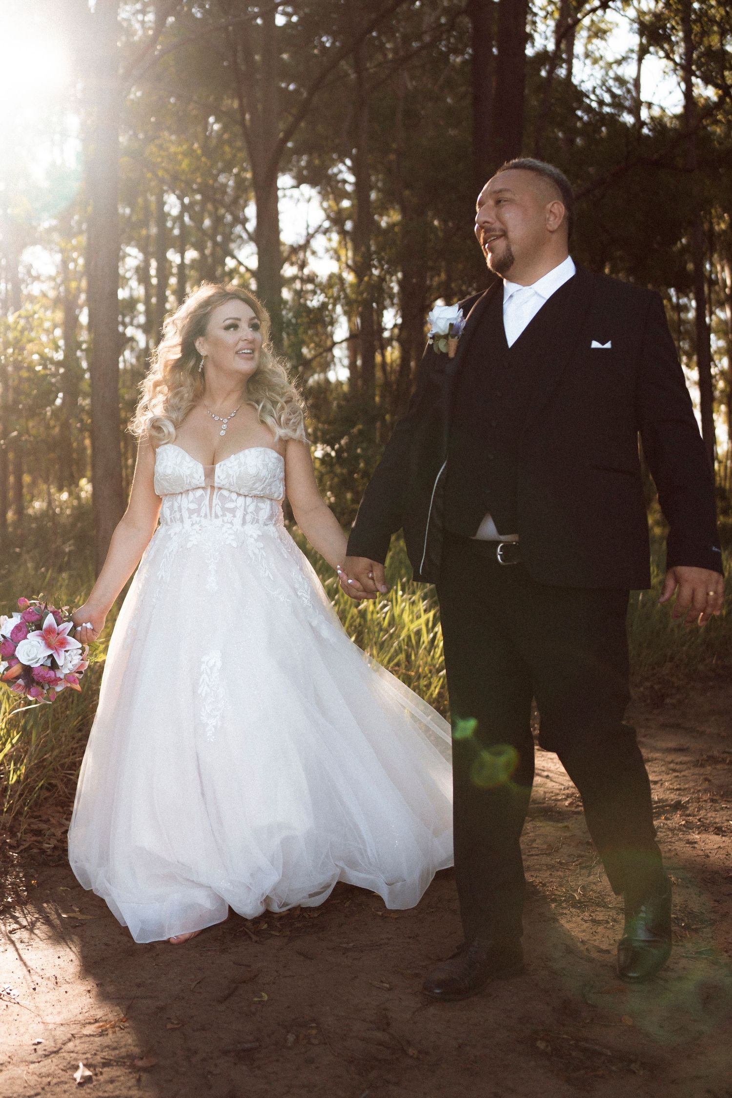 Bride in white ball gown and groom in black tuxedo walk hand in hand through sunlit forest on their wedding day.