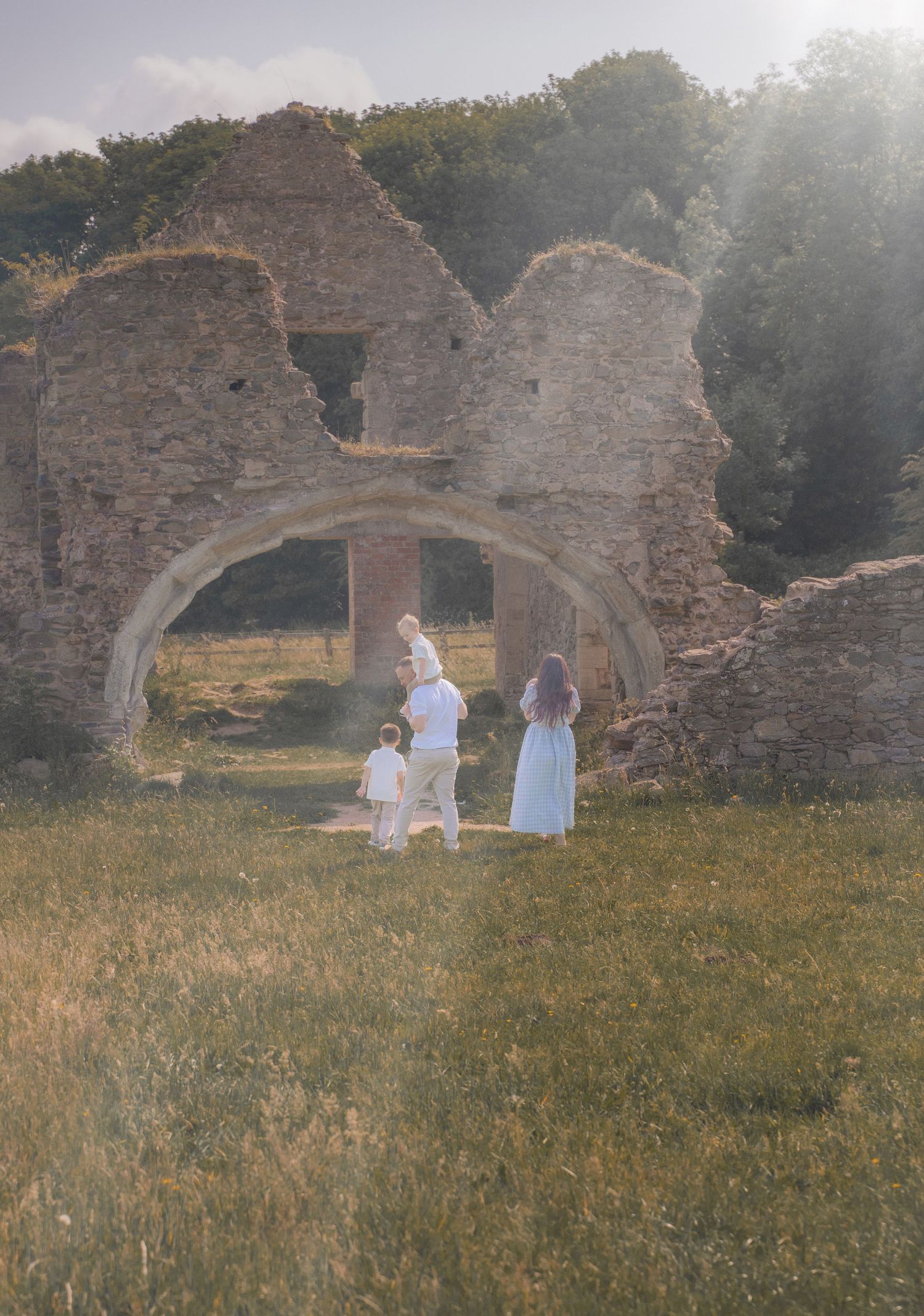 A family of four walking hand in hand beneath a stone archway at Grace Dieu Priory near Coalville, with soft evening light filtering through