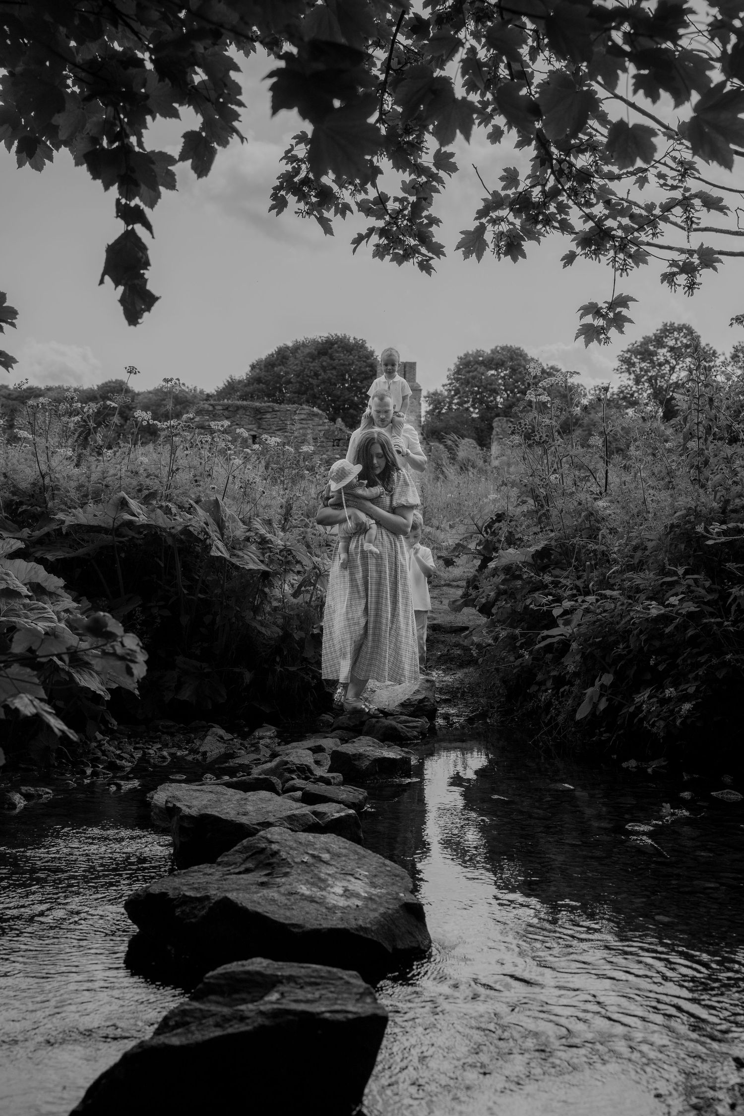 A mother carrying her baby while walking barefoot along a stream surrounded by wild greenery near Coalville. A quiet, natural moment during