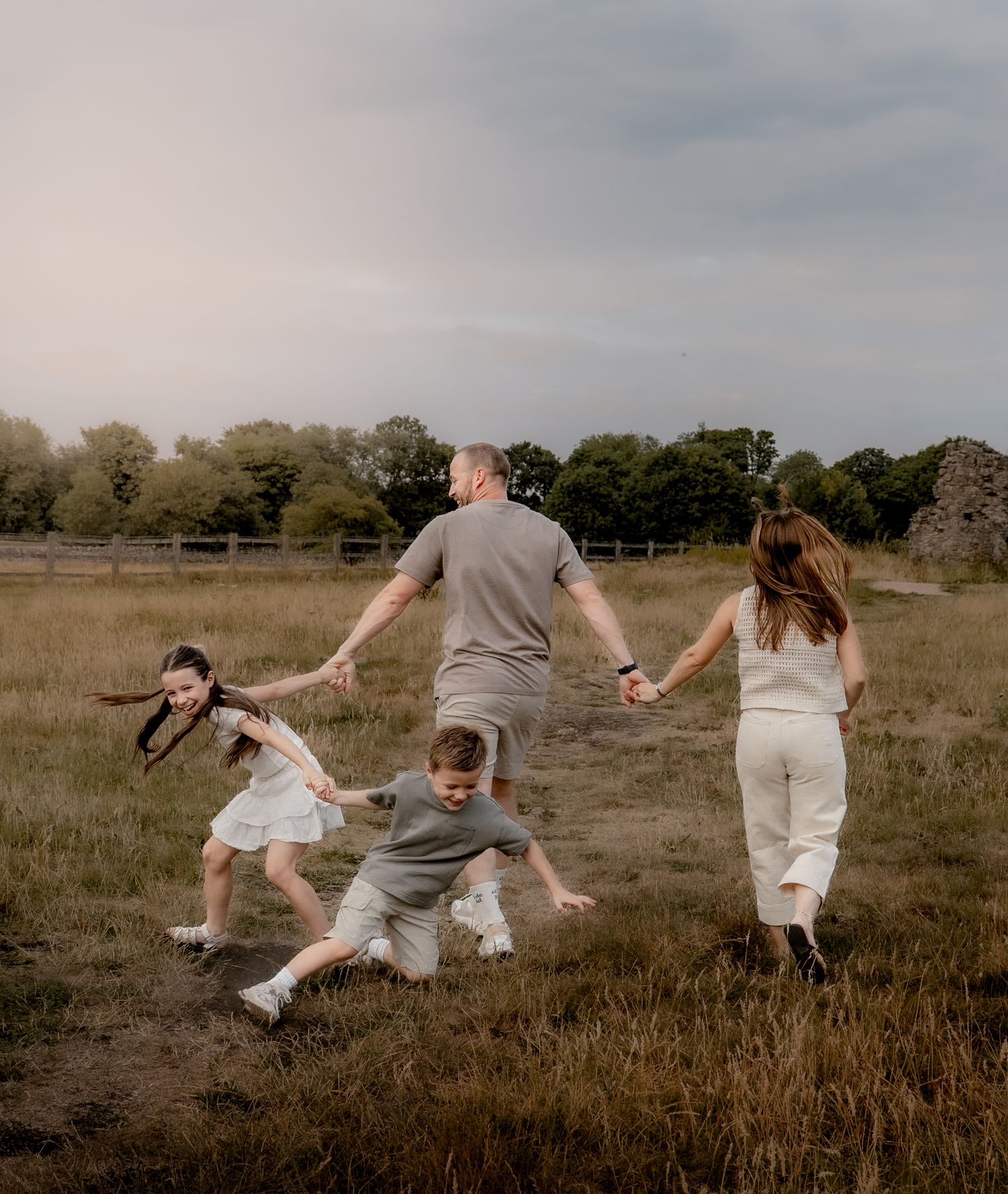 Parents and children running hand-in-hand through long grass at sunset during a relaxed family photo shoot in Derbyshire. Captured mid-laugh