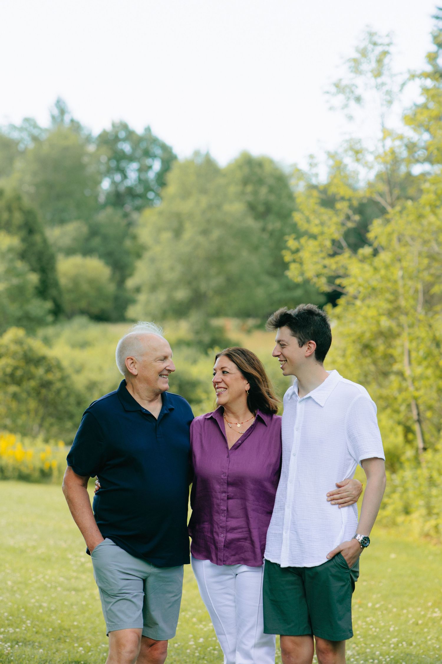 Three people walk together and share a laugh in a sunny meadow with yellow wildflowers and green trees in the background.