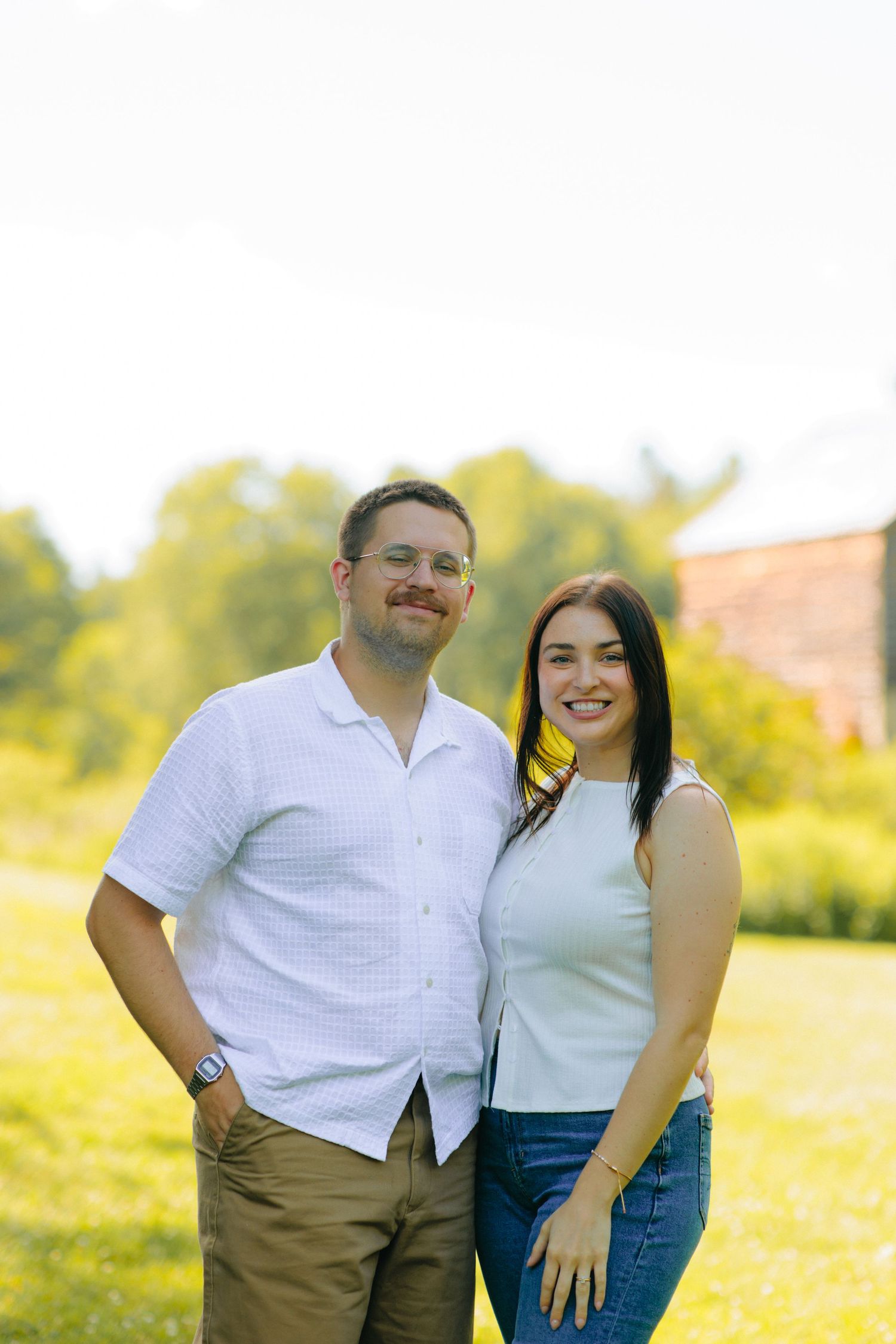 A couple in casual attire poses together on a sunny day with green foliage and trees in the background.