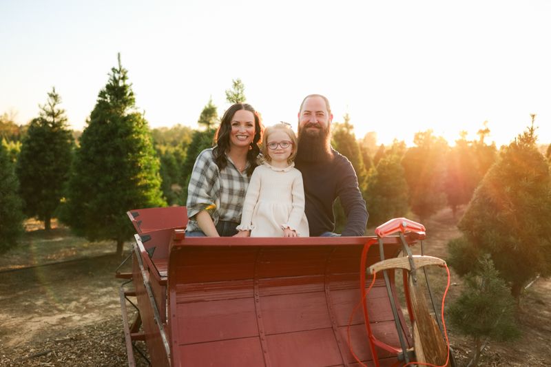 Family poses together in back of vintage red truck at Christmas tree farm during golden hour.