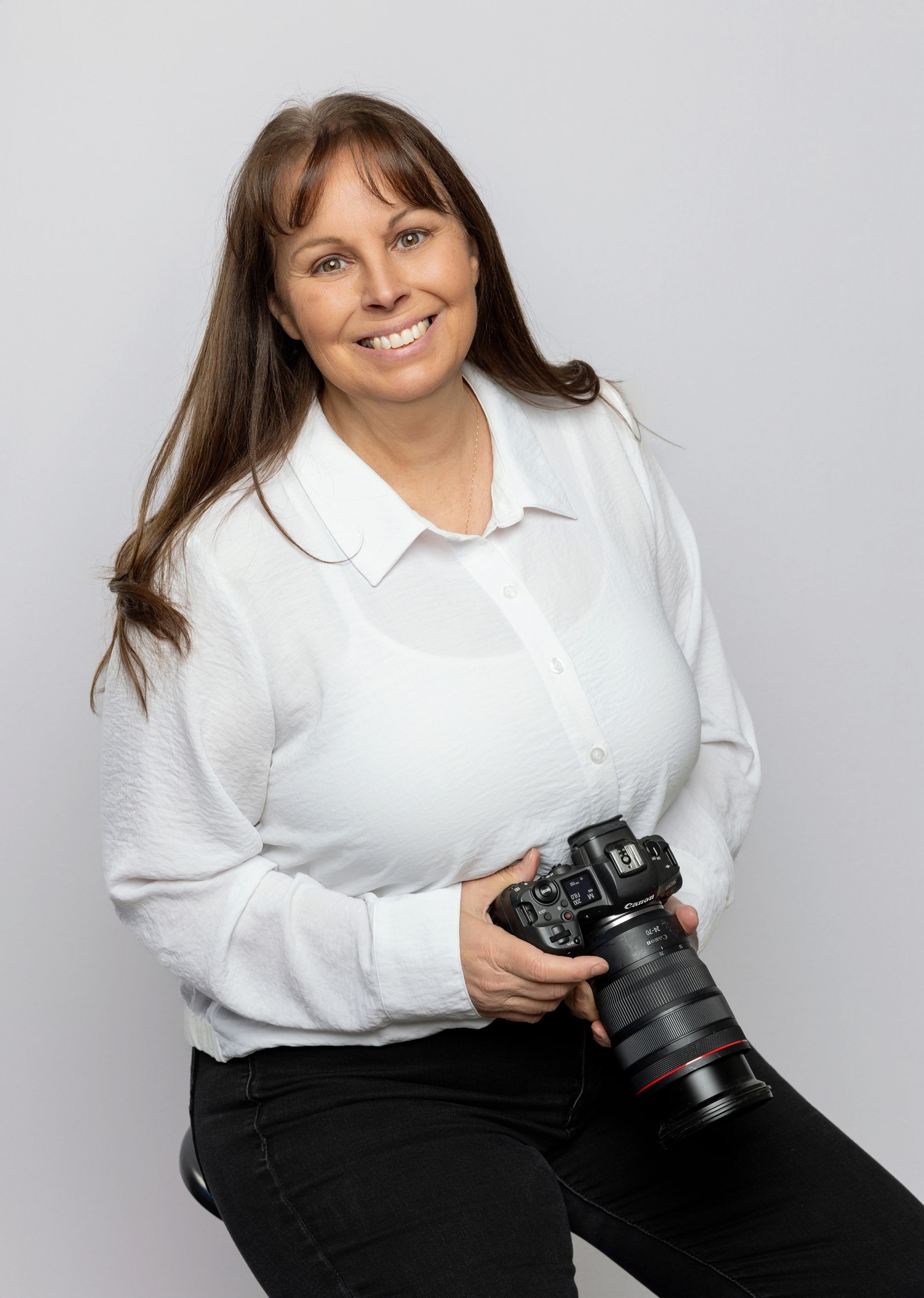 Professional photographer in white blouse poses with camera against studio backdrop.