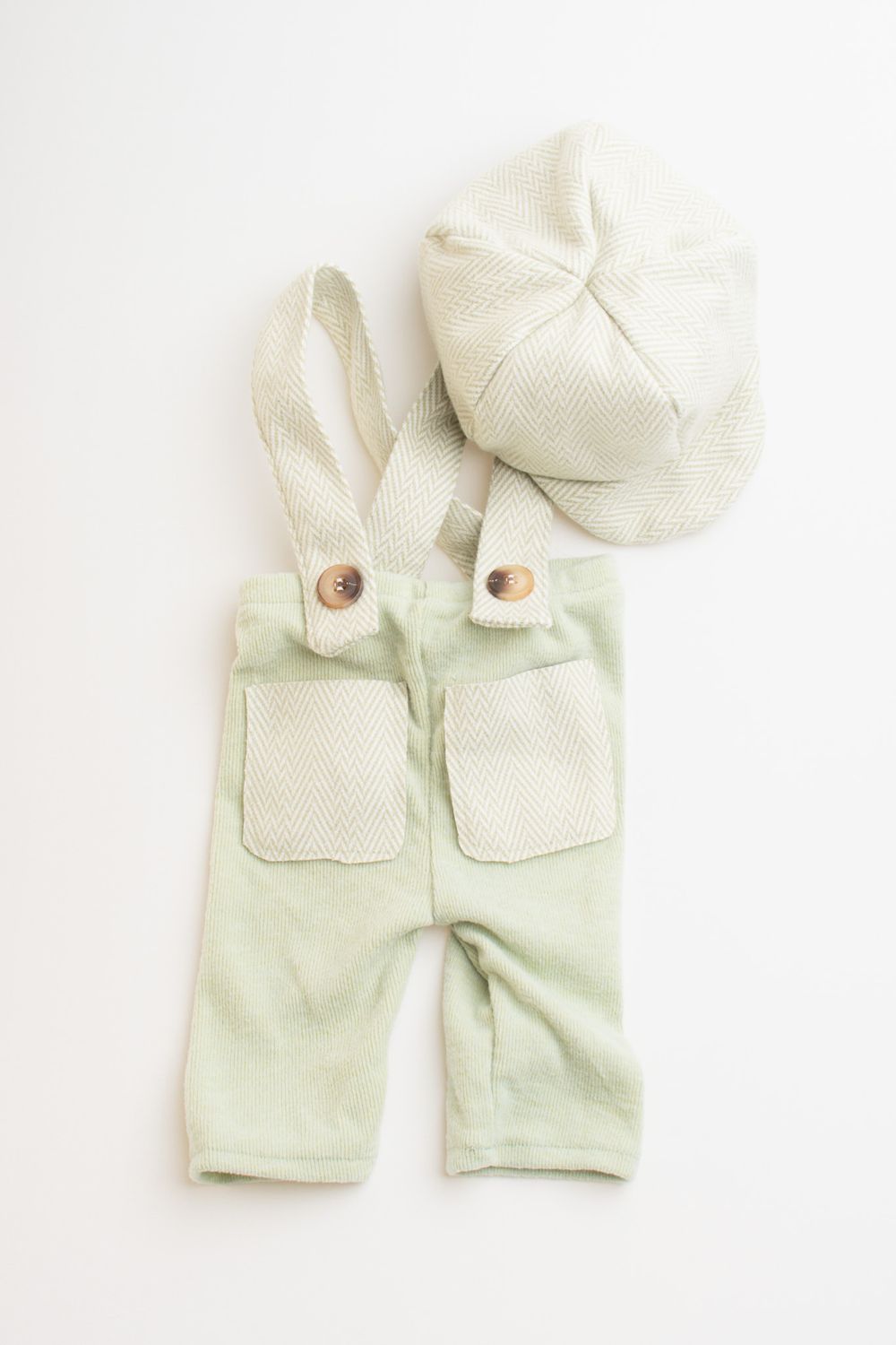 Mint green baby overalls with front pockets and white newsboy cap displayed on white background.