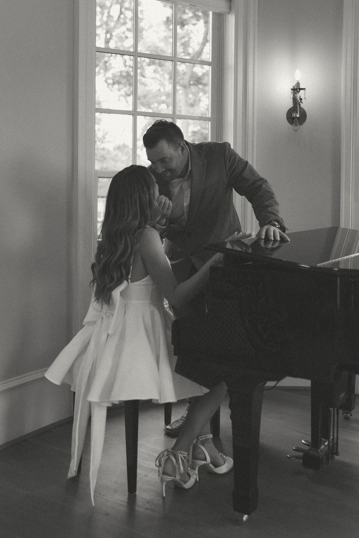 A romantic black and white photograph of a couple sharing an intimate moment by a piano in sunlit room.