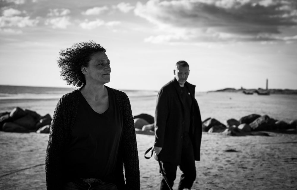 Schwarz-Weiß-Foto eines Paares am Strand in Dänemark. Beide laufen Hand in Hand am Meer entlang, sanfte Wellen, ruhige und intime Atmosphäre