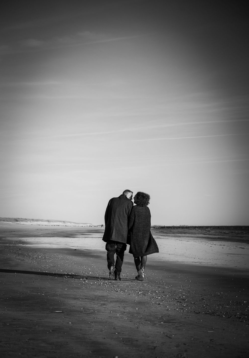 Schwarz-Weiß-Foto eines Paares am Strand in Dänemark. Beide laufen Hand in Hand am Meer entlang, von hinten gesehen, weg vom Betrachter.